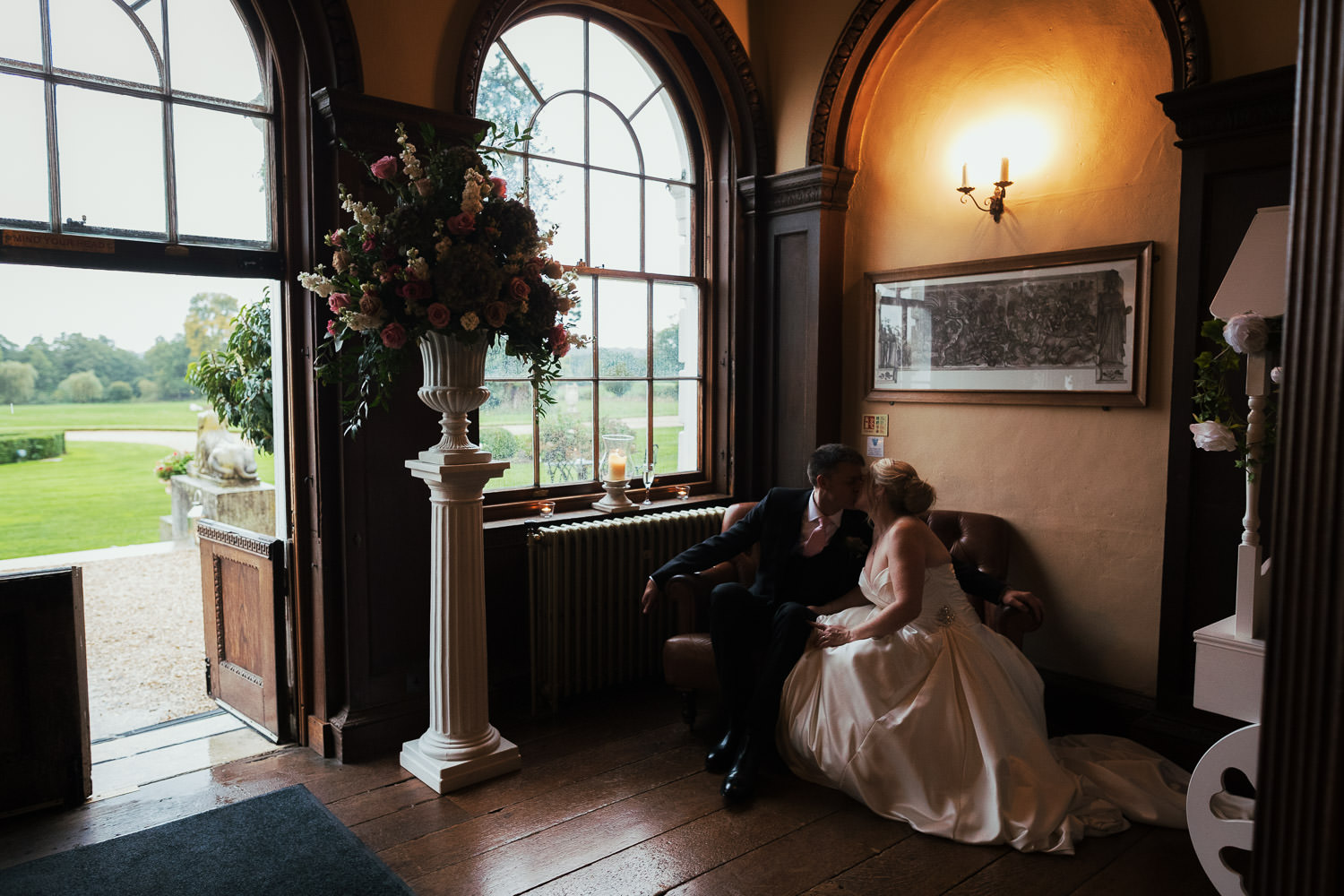 Newly weds kiss while sitting in a sofa in the library at Gosfield Hall. There are windows and a door on the left of the picture.