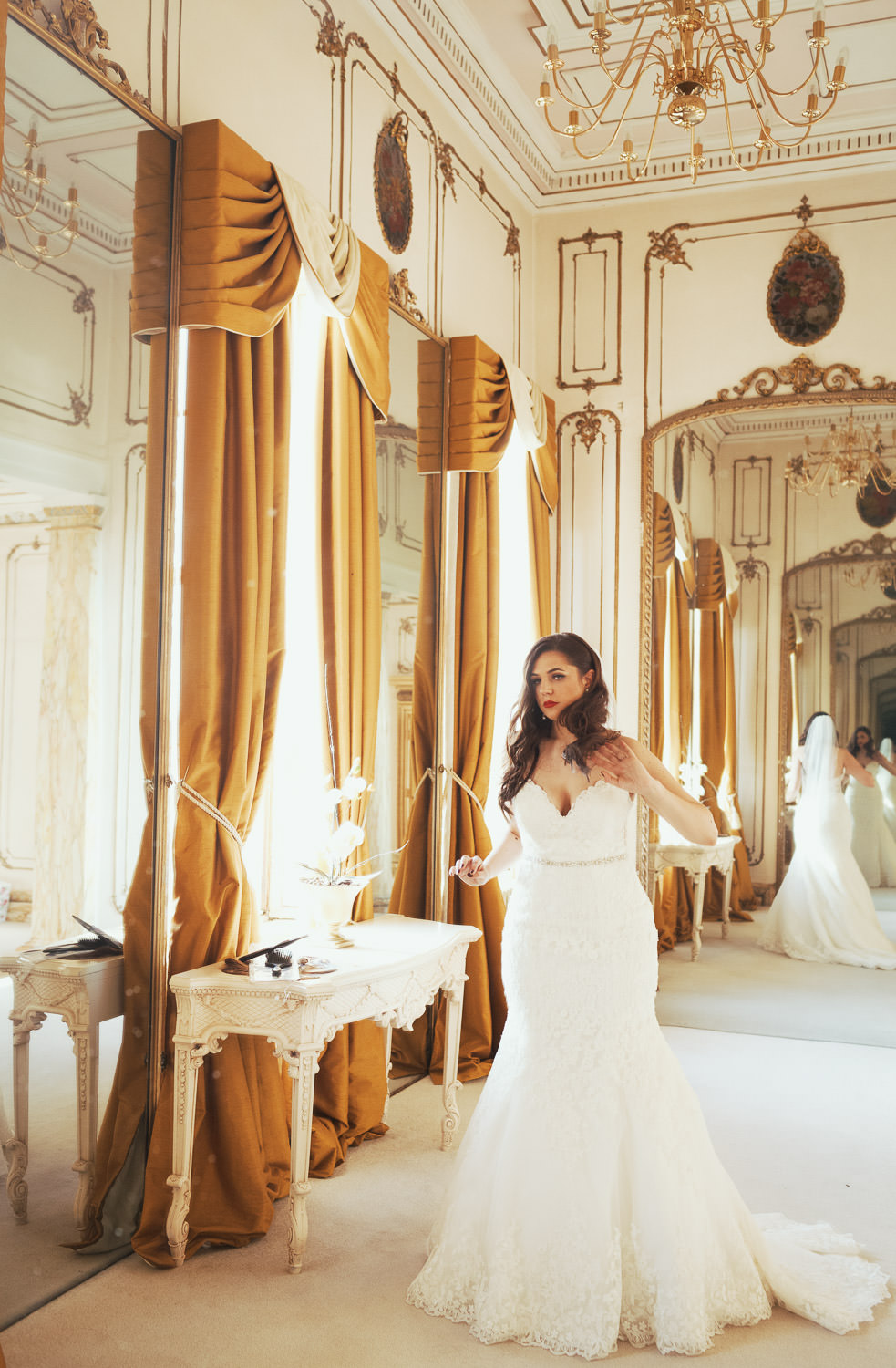 A bride in a long white dress adjusting her hair. Mirrors behind and in front of her. In the Rococo Bridal suite (Late Baroque) bridal room at Gosfield Hall.