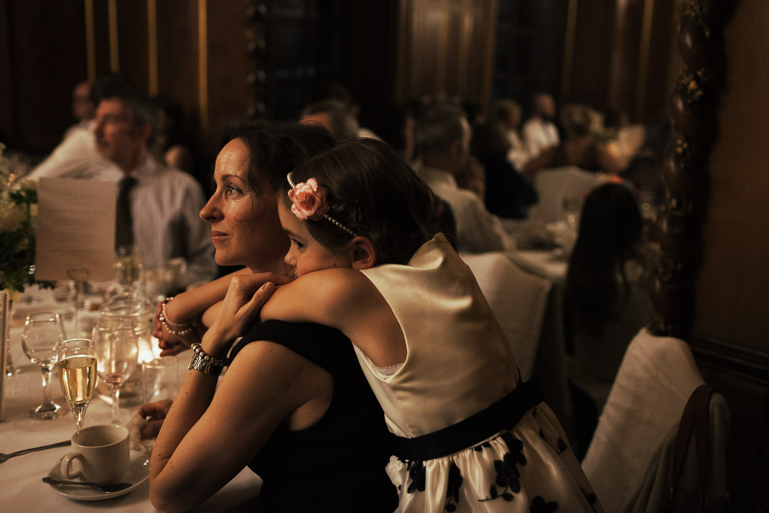 A young girl hugging her mum over her shoulders. Her mum is sat at a round table during a wedding breakfast. In the ballroom at Gosfield Hall.