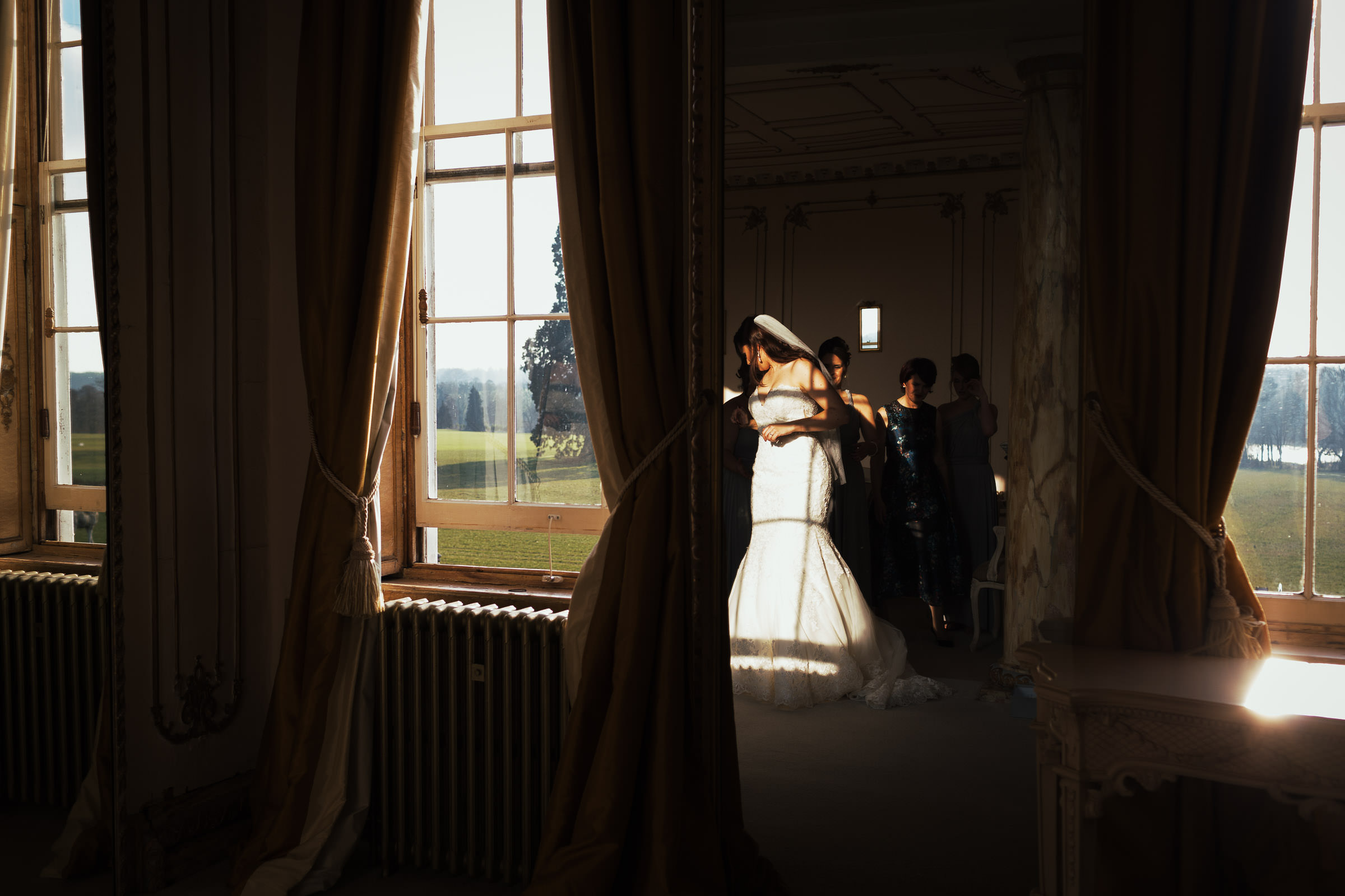 In the bridal suite at Gosfield Hall a bride is getting in her dress with help from her mum and bridesmaids. Her reflection is in the mirror, you can see the grounds outside through the windows. A real wedding moment.