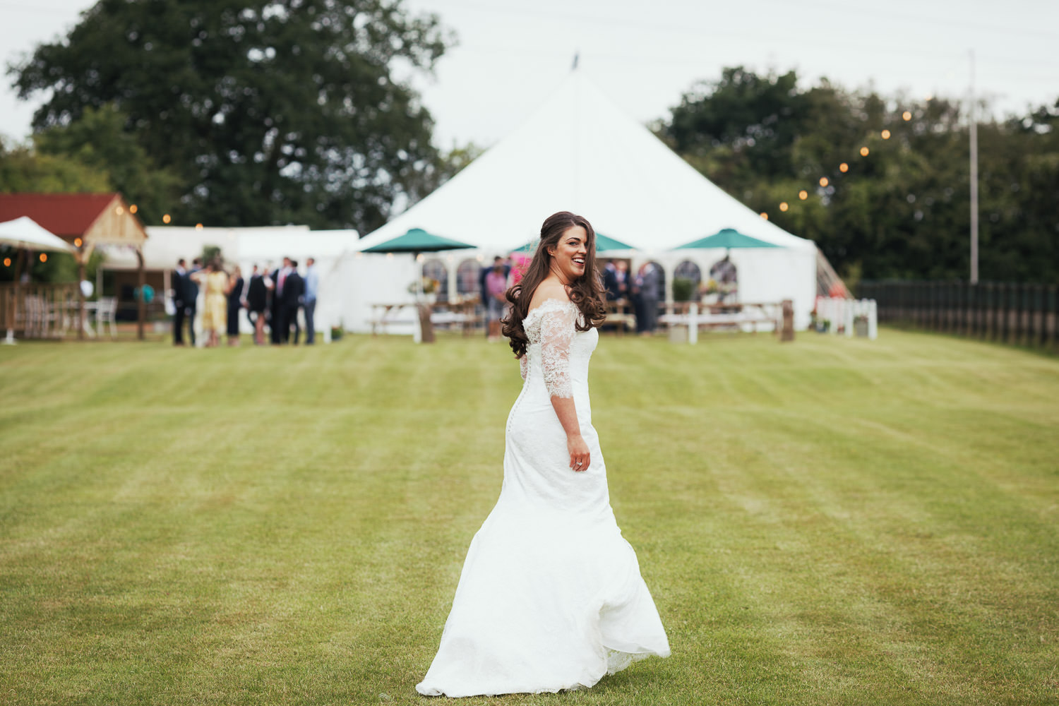 Essex wedding photography testimonial picture. The bride is in a field looking back at the camera and smiling. A wedding marquee is in the distance.