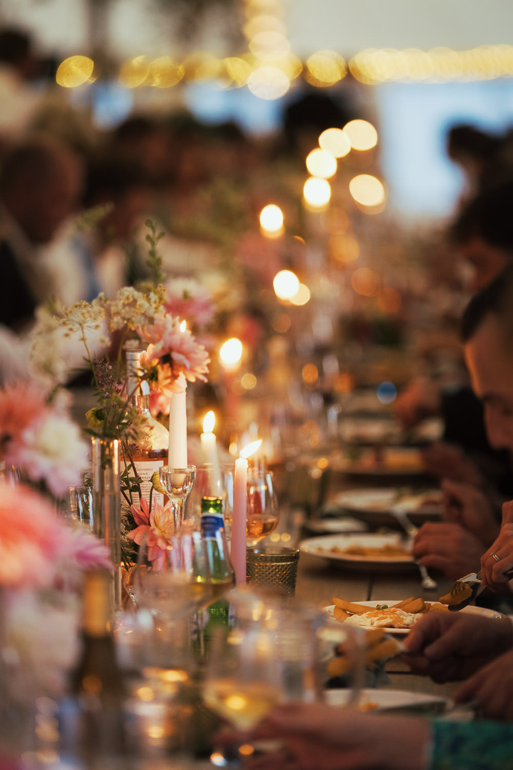 A row of people on a long table eating ham, egg, and chips from Anna Shutes Catering. The table is decorated with flowers and candles.