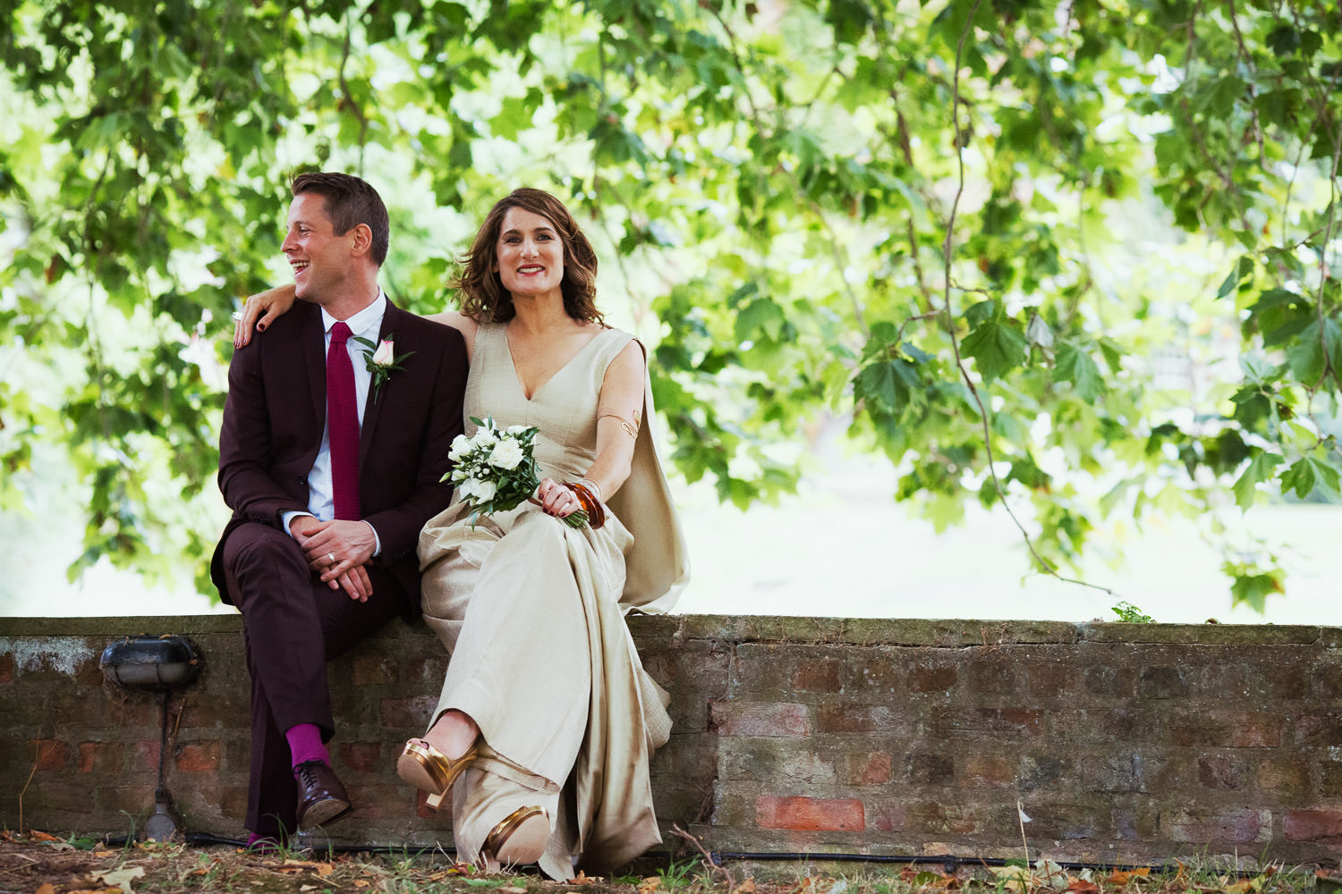 Just married couple sat on a wall by the River Cam at St John's College. The leaves are green. Man's suit is aubergine and bride is wearing a golden jumpsuit and cape.
