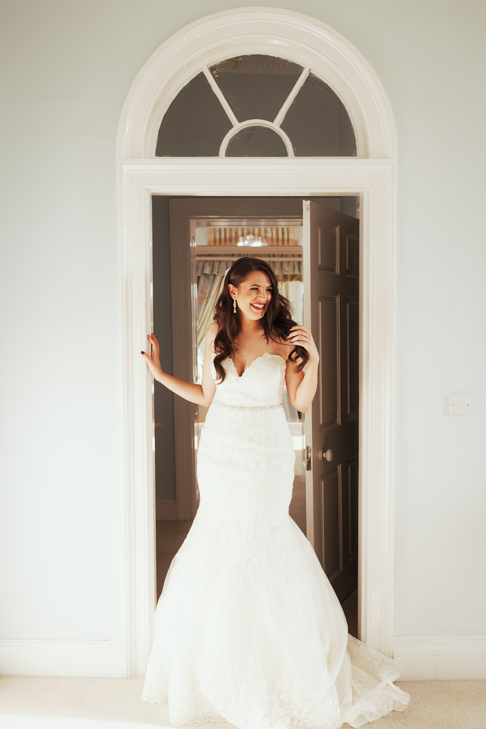Bride in lace mermaid style wedding dress by Mori Lee, style 2713. Long dark hair and red lipstick, smiling in doorway of Queen's Apartment. Captured by regular Gosfield Hall documentary wedding photographer Tracy, based in Essex.