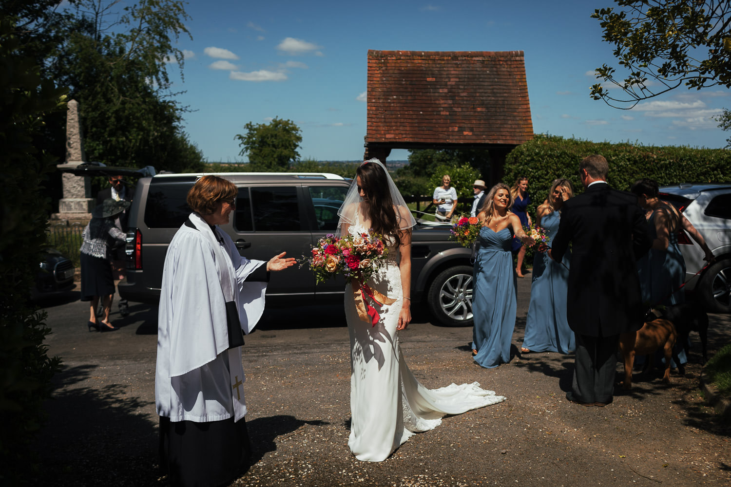 Reverend Julie Willmot greets a bride as she arrives at All Saints Church in Purleigh Essex, near Maldon. Bridesmaids are behind her in blue dresses and the sky is blue. Bride is wearing Stella York 6648 from The Wedding Shop in Colchester. Photography by South Woodham Essex wedding photographer Tracy.