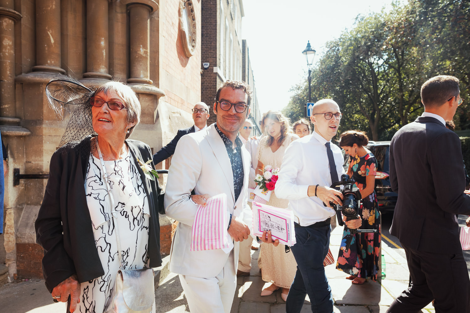 Jake Maskall escorting another guest out of the Union Chapel after a wedding ceremony. He is wearing a white suit, the woman is wearing a black and white dress and a black fascinator.