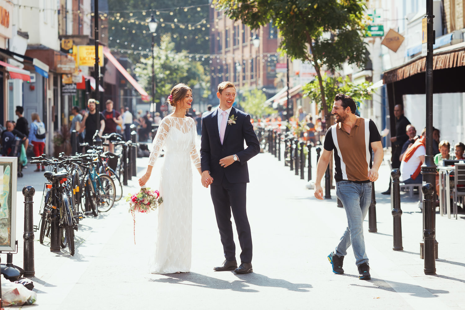 Couple posing for a portrait in the Exmouth Market on their wedding day. A random stranger is smiling at them after congratulating them. Then they kiss. The bride is wearing the Willow Halfpenny dress. A stranger says hello.