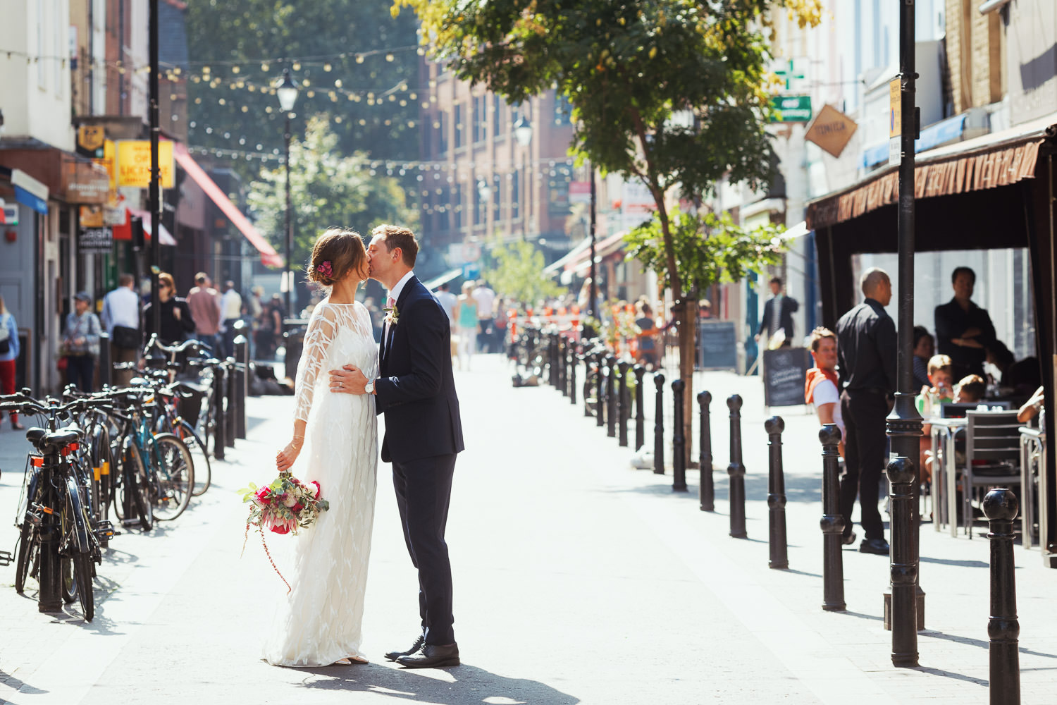 Couple posing for a portrait kiss in the Exmouth Market on their wedding day. A random stranger is smiling at them after congratulating them. Then they kiss. The bride is wearing the Willow Halfpenny dress.