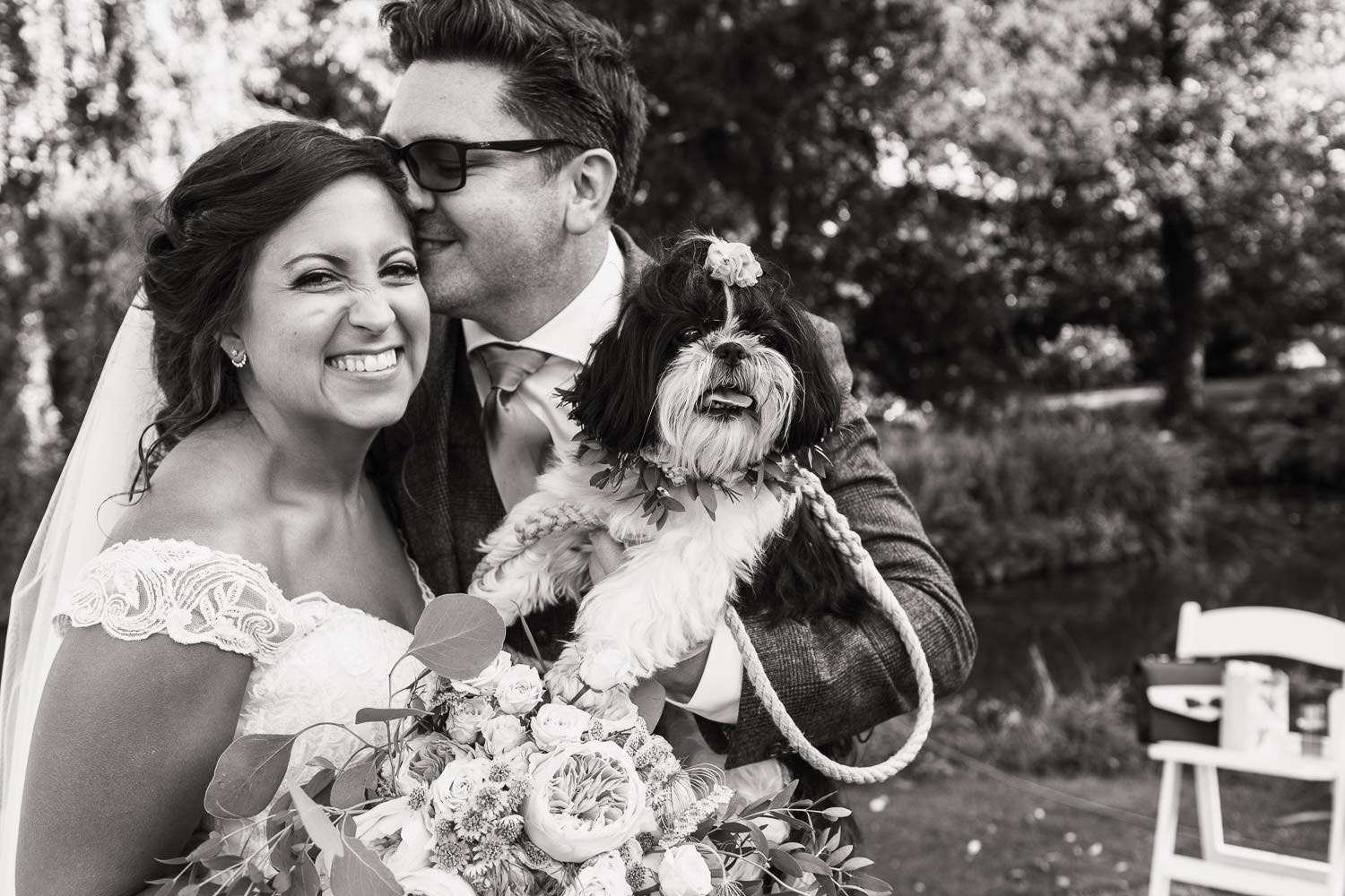 A black and white portrait of a bride, groom and their dog. The man is pressing his nose up to his new wife's head. She is smiling at the camera.