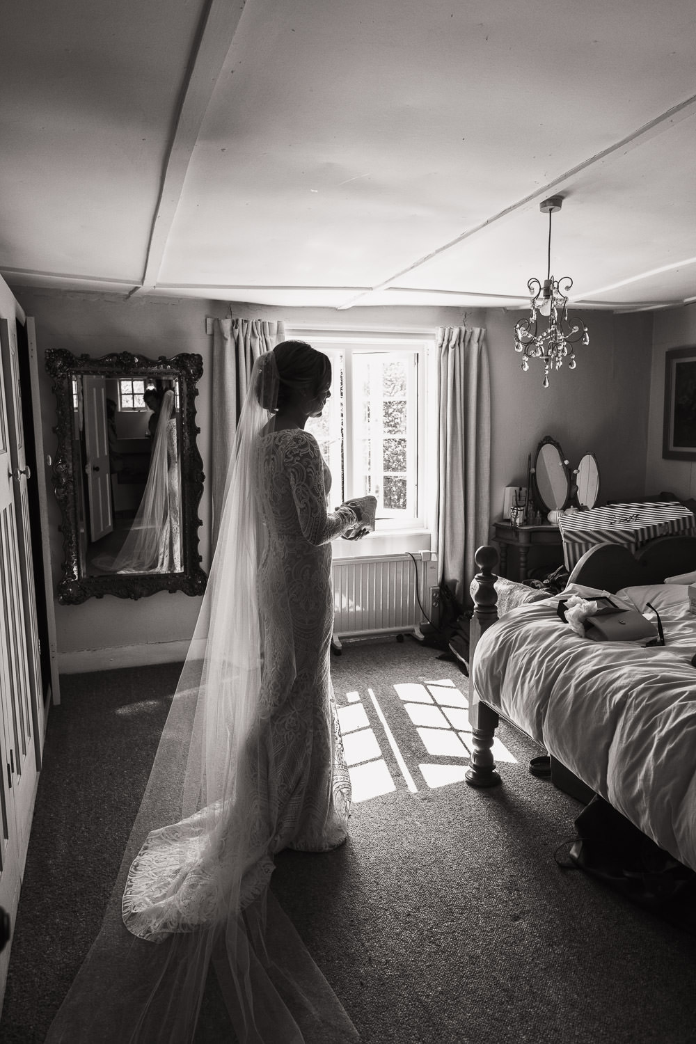 Bride in full length lace dress and veil stands in bedroom with sunlight streaming through window, holding purse. Custom-made by Louise Sullivan of Louise Bridal, Stow Maries near South Woodham.