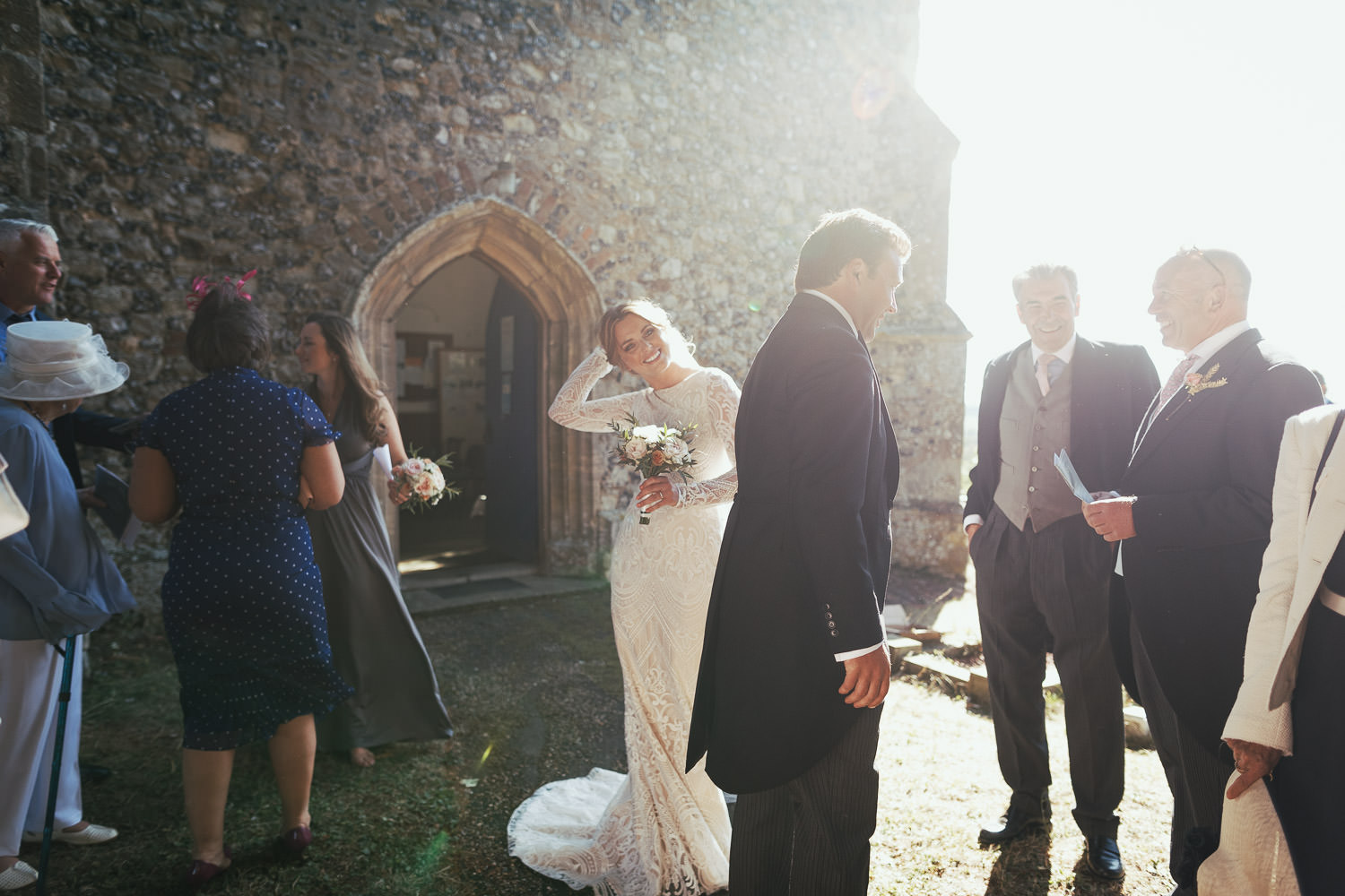Wedding guests are chatting to each other but the bride is looking at the camera, illuminated by the sunlight. The door of The Church of St Mary & St Margaret in Stow Maries is behind her. She is wearing a long lace wedding dress with long sleeves.