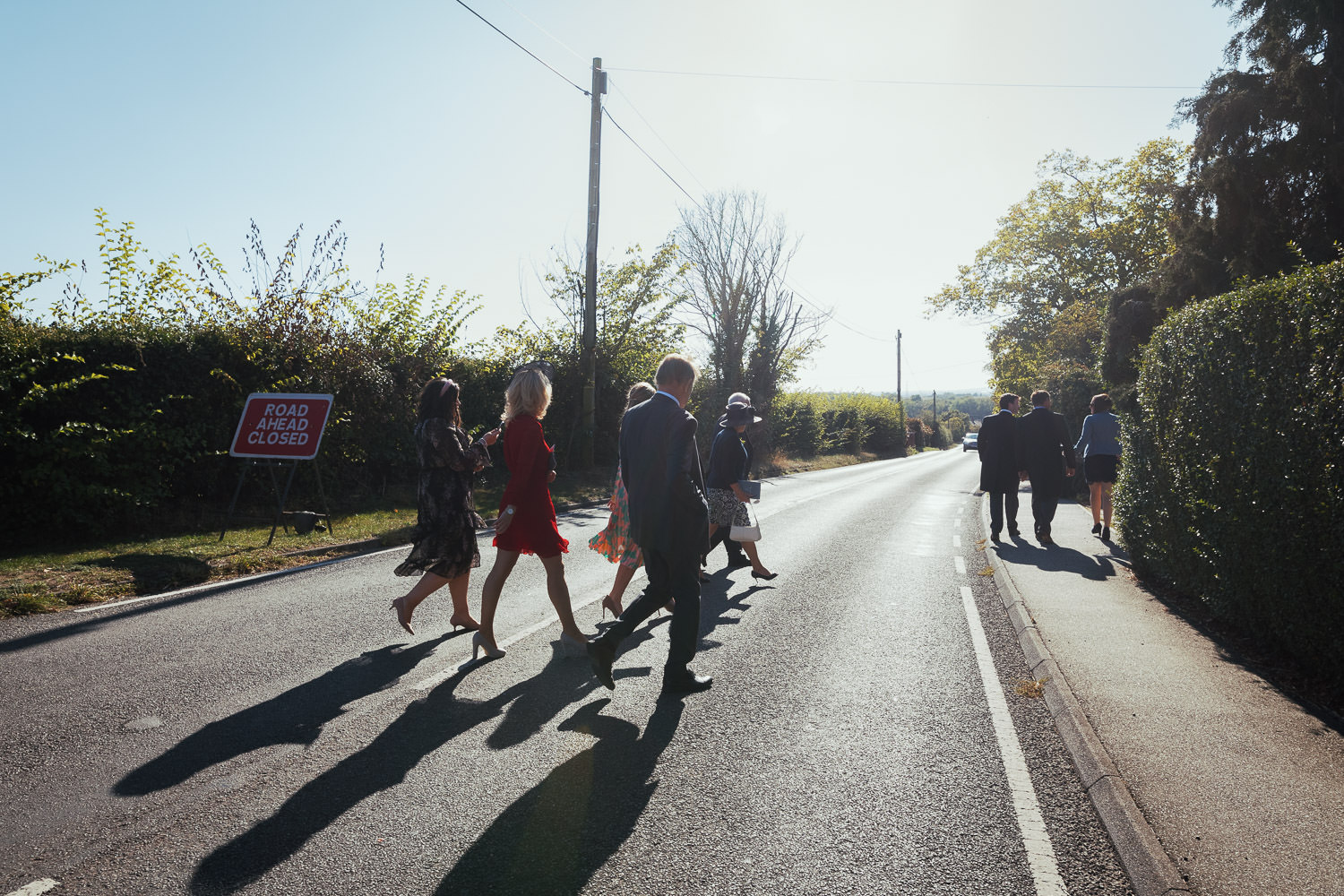 People leaving the church after a wedding walk across Woodham Road in Stow Maries. It's a sunny day and they cast shadows on the road.