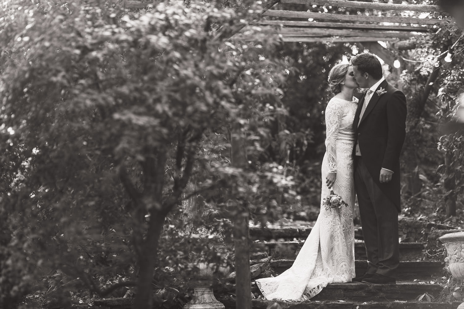 On the steps under a pergola in a family garden a bride and groom kiss. They are surrounded by trees and plants. The bride is wearing a long lace wedding dress with long sleeves. The groom is in a morning suit with a waistcoat.
