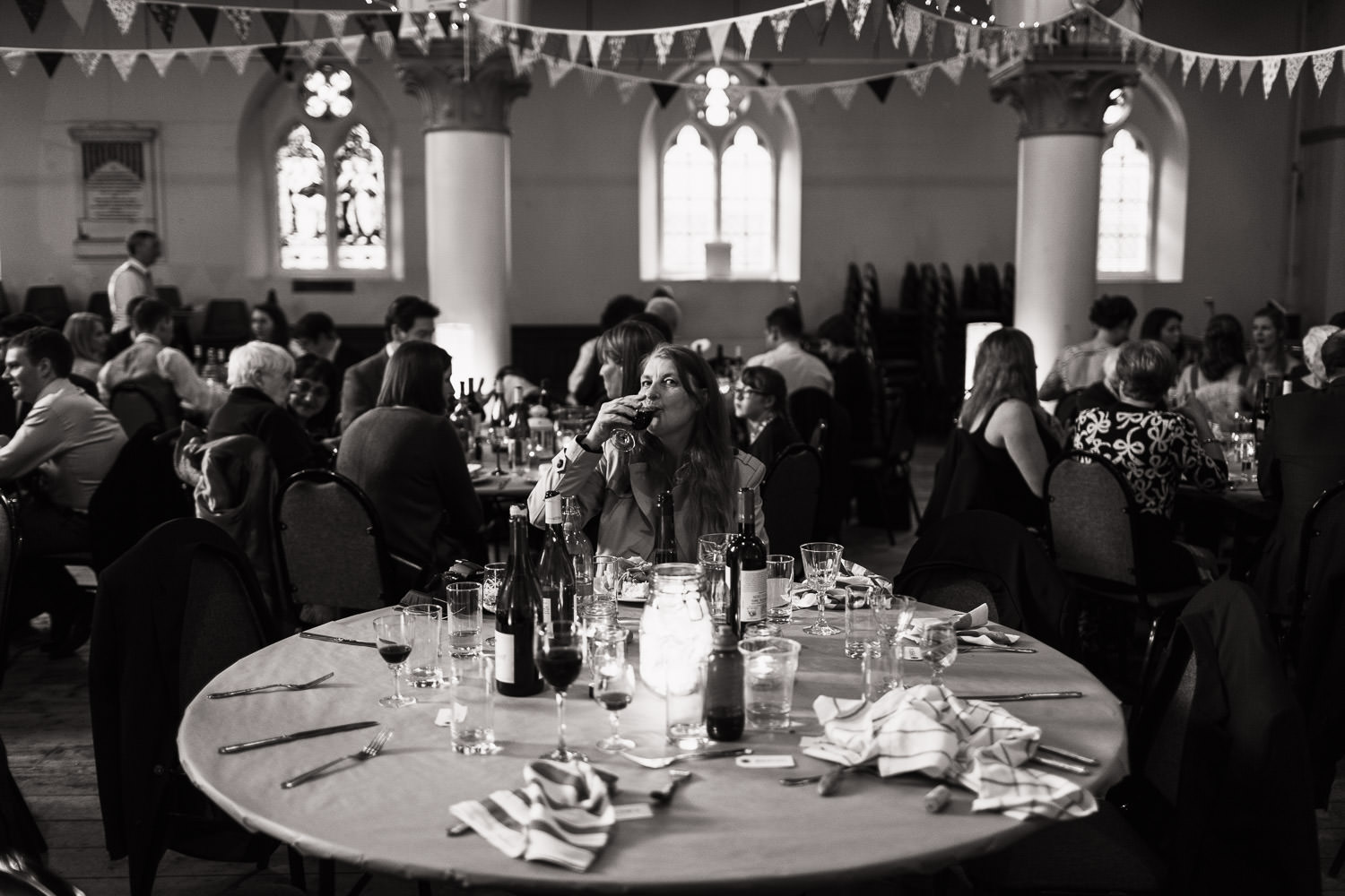 A woman is sat alone at a round table while the other people get their food. She's looking at the camera and drinking a glass of wine. A reportage style unposed wedding photograph. At the Elmgrove Centre.