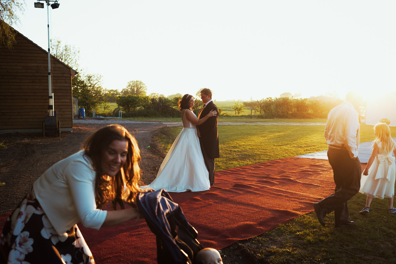 A candid moment from an Essex wedding. The bride and groom or stood facing each other while wedding guests walk past. A woman is pushing a buggy in the foreground. The sun is setting.