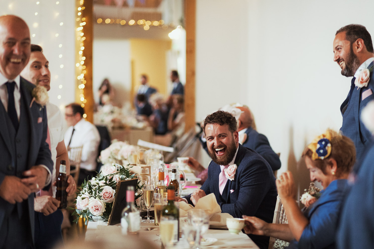 A groom sat at the top table, surrounded by guests and laughing. At Houchins venue in Essex.