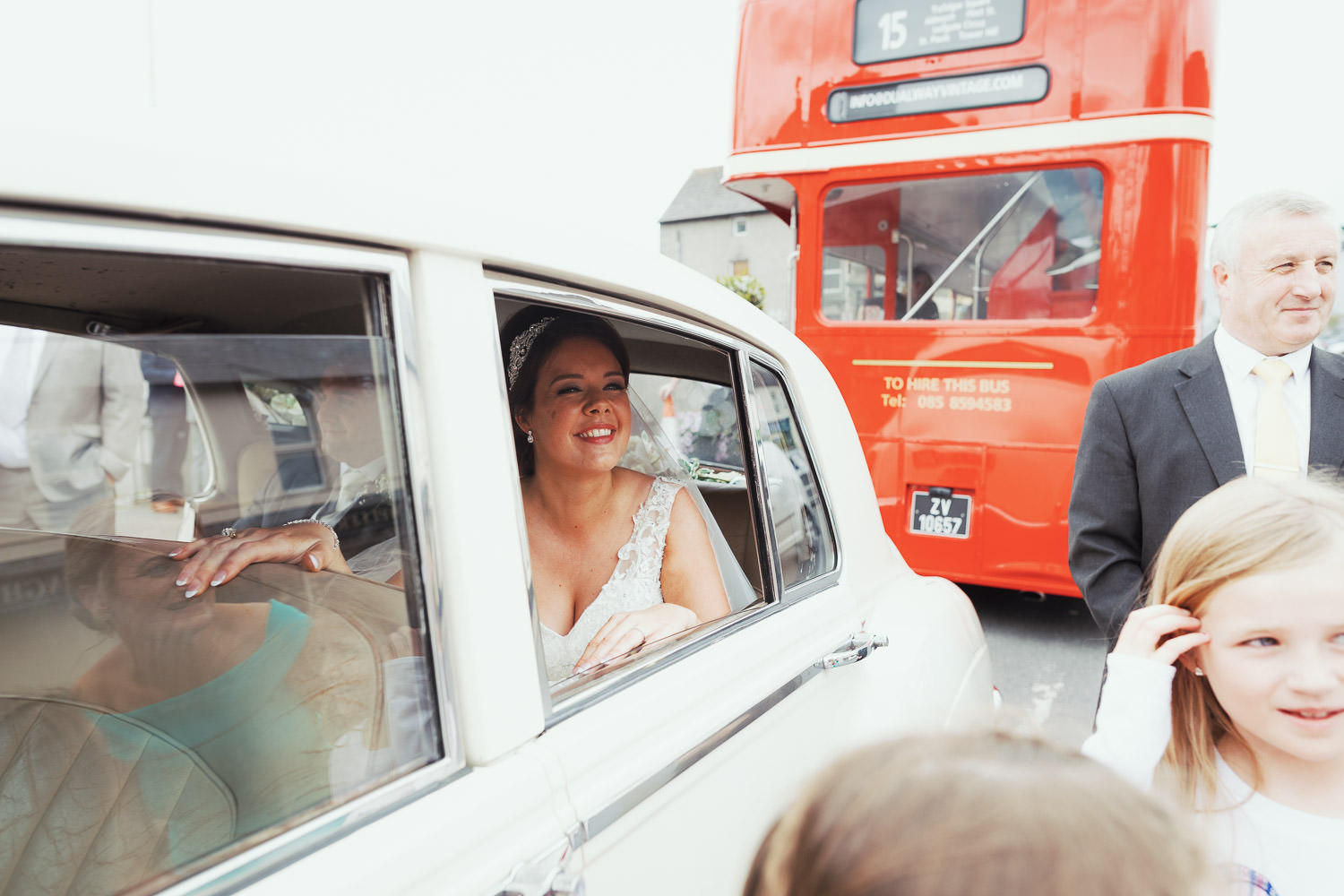 Bride looks out of the car window with a smile on her face, a young girl is putting her hair behind her ear.