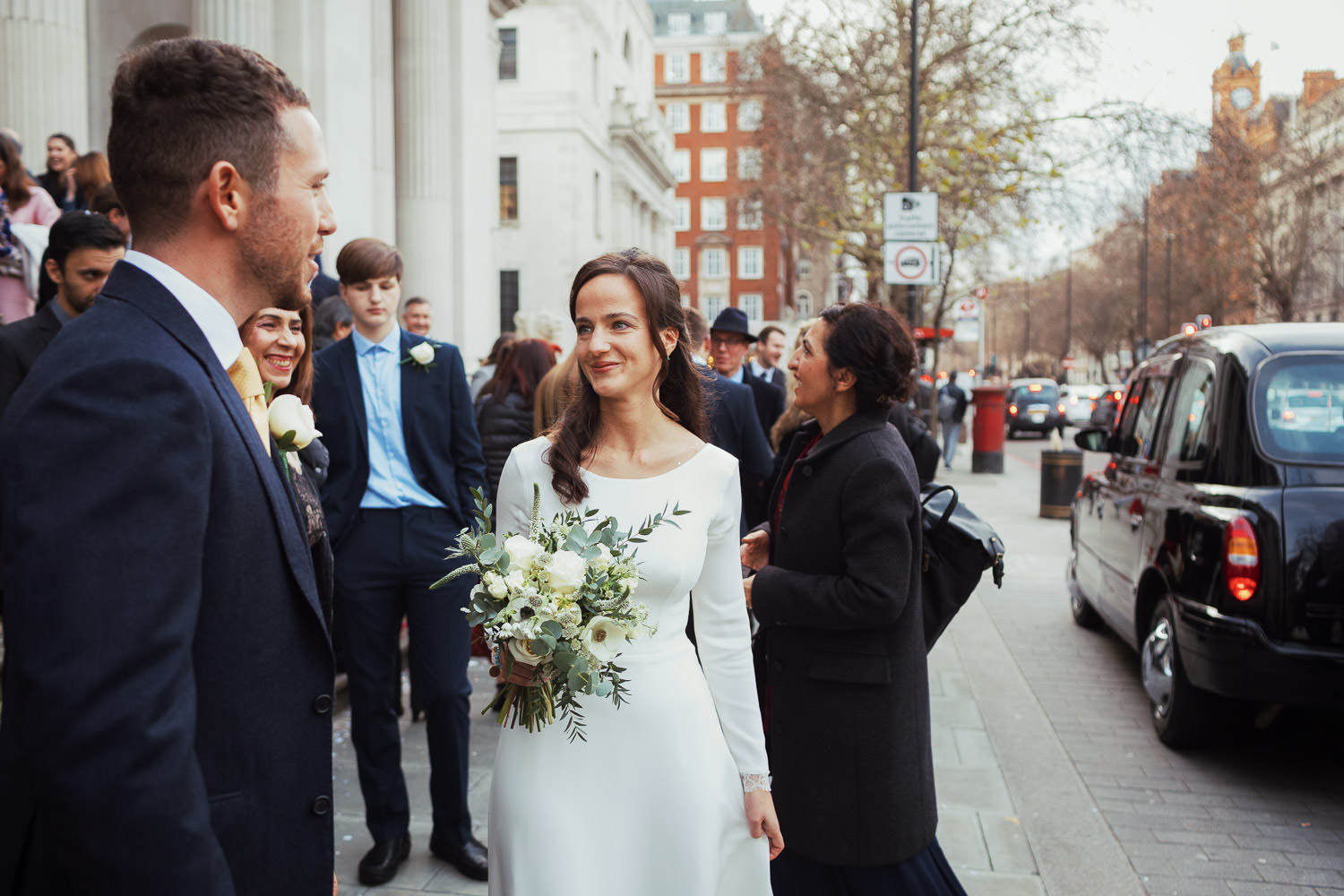 A reportage style portrait of a bride, unposed, at a wedding. She is holding white flowers, wearing a long white dress and is smiling, She is stood outside the Old Marylebone Town Hall after the ceremony. Her husband is in shot, she is surrounded by wedding guests as black cabs turn up.