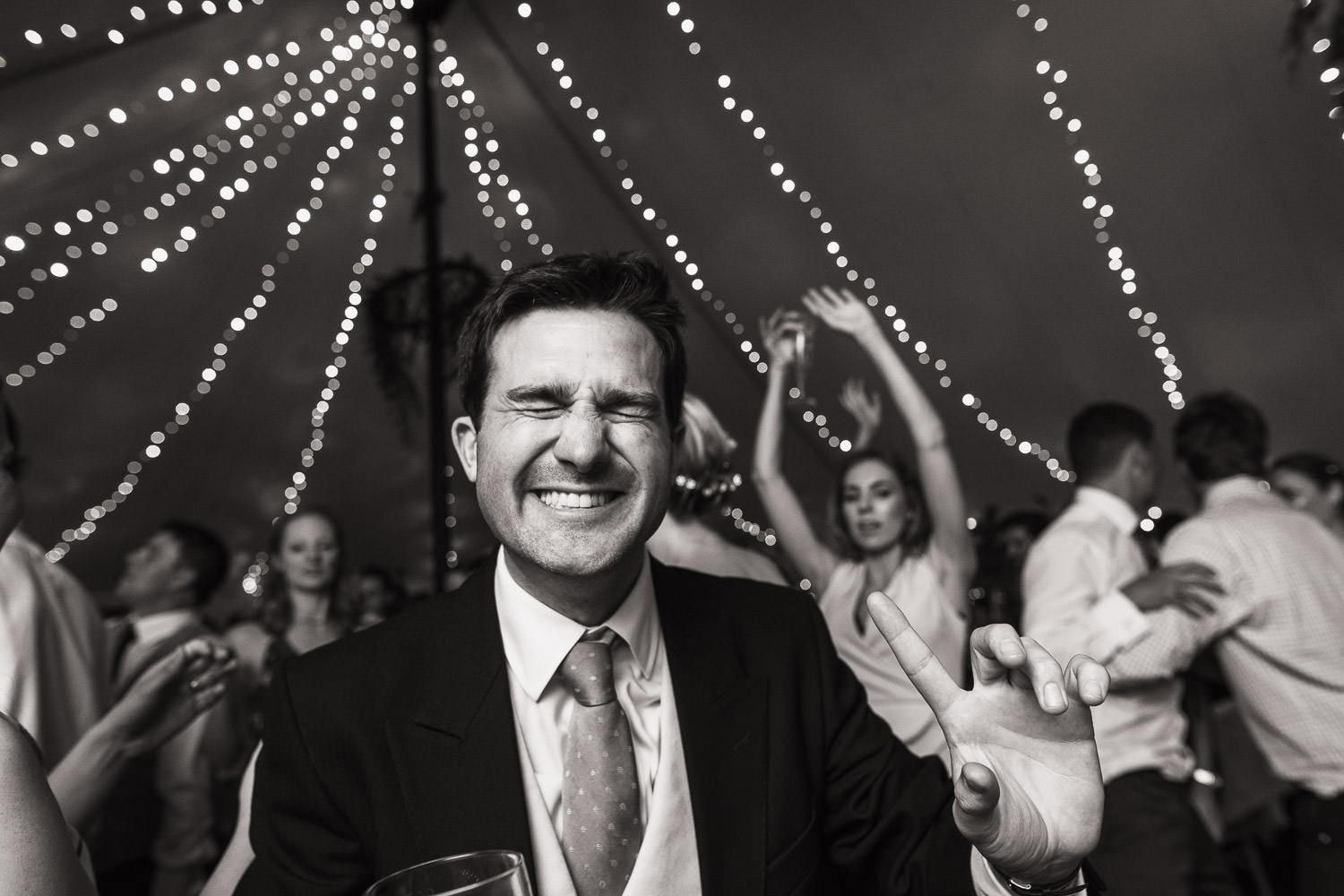 Man closing his eyes and point as people dance behind him at a wedding in Essex.