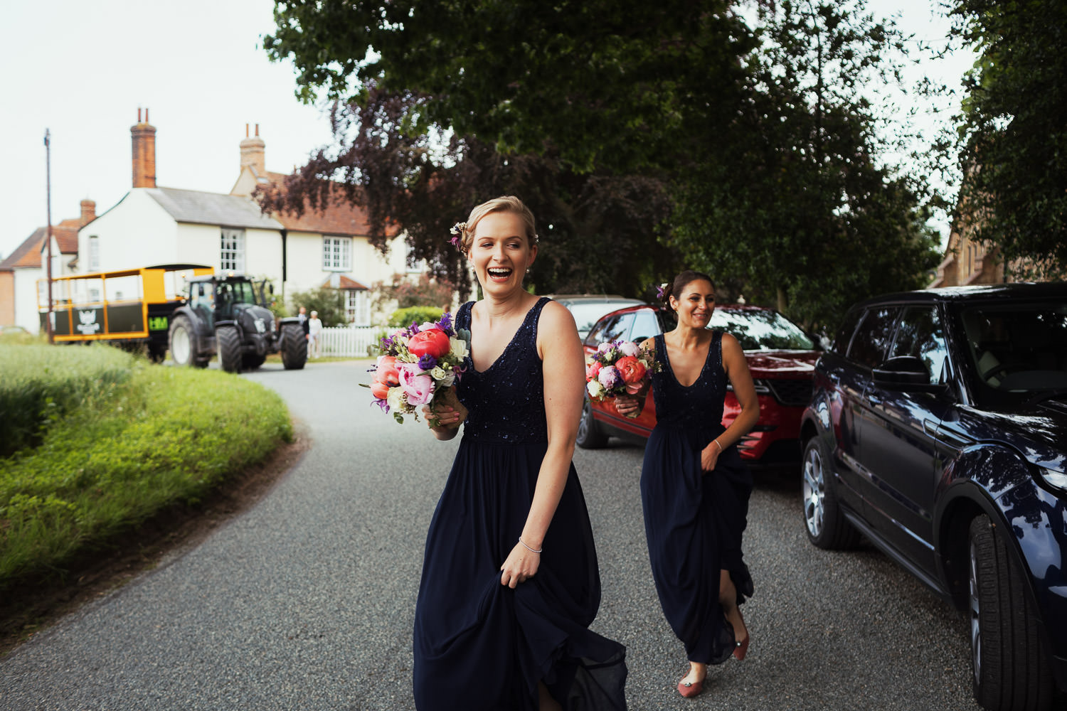 Two bridesmaids cross the road outside St Peter's Church in Great Totham. A blonde bridesmaid is laughing, they are wearing navy blue long dresses.