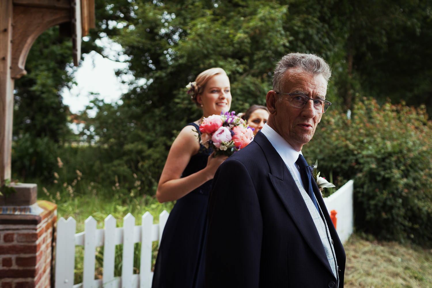 Man in a suit looks at the camera. A bridesmaid in the background.