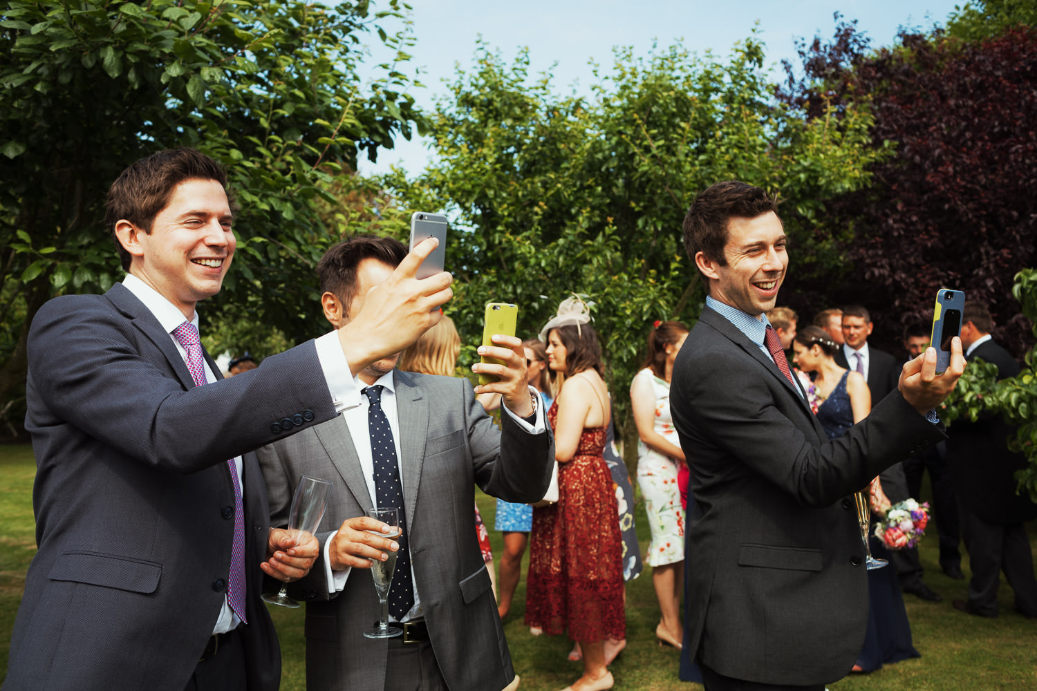 Men taking photos with their phones in a garden at a wedding reception.
