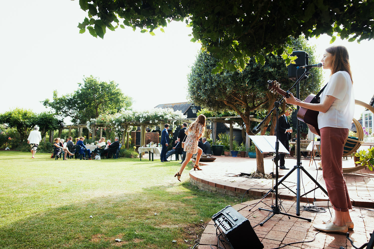A woman is playing guitar and singing to a garden full of people enjoying drinks after a wedding.