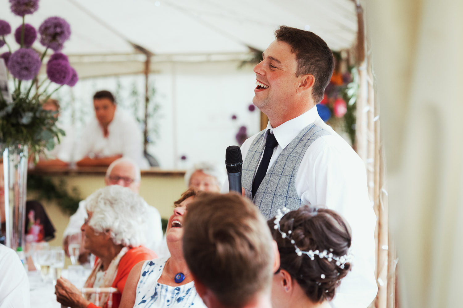 Groom giving a speech at a wedding breakfast in a white marquee. People are sitting at tables.