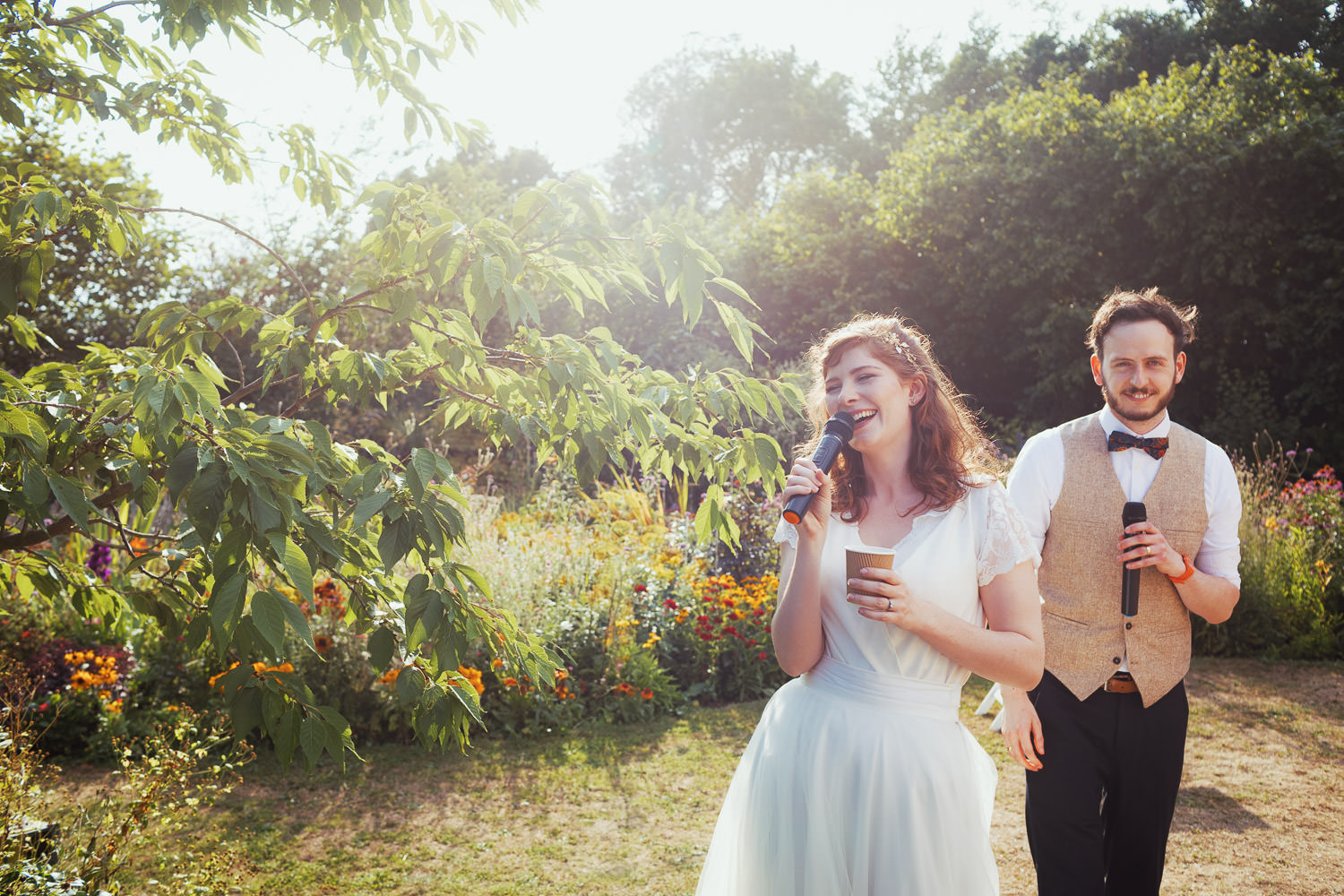 Woman in her homemade wedding dress holds a cup of tea and a microphone, she is giving a speech at her wedding. Her husband is stood just behind her. They are in a sunny garden full of yellow and red flowers.