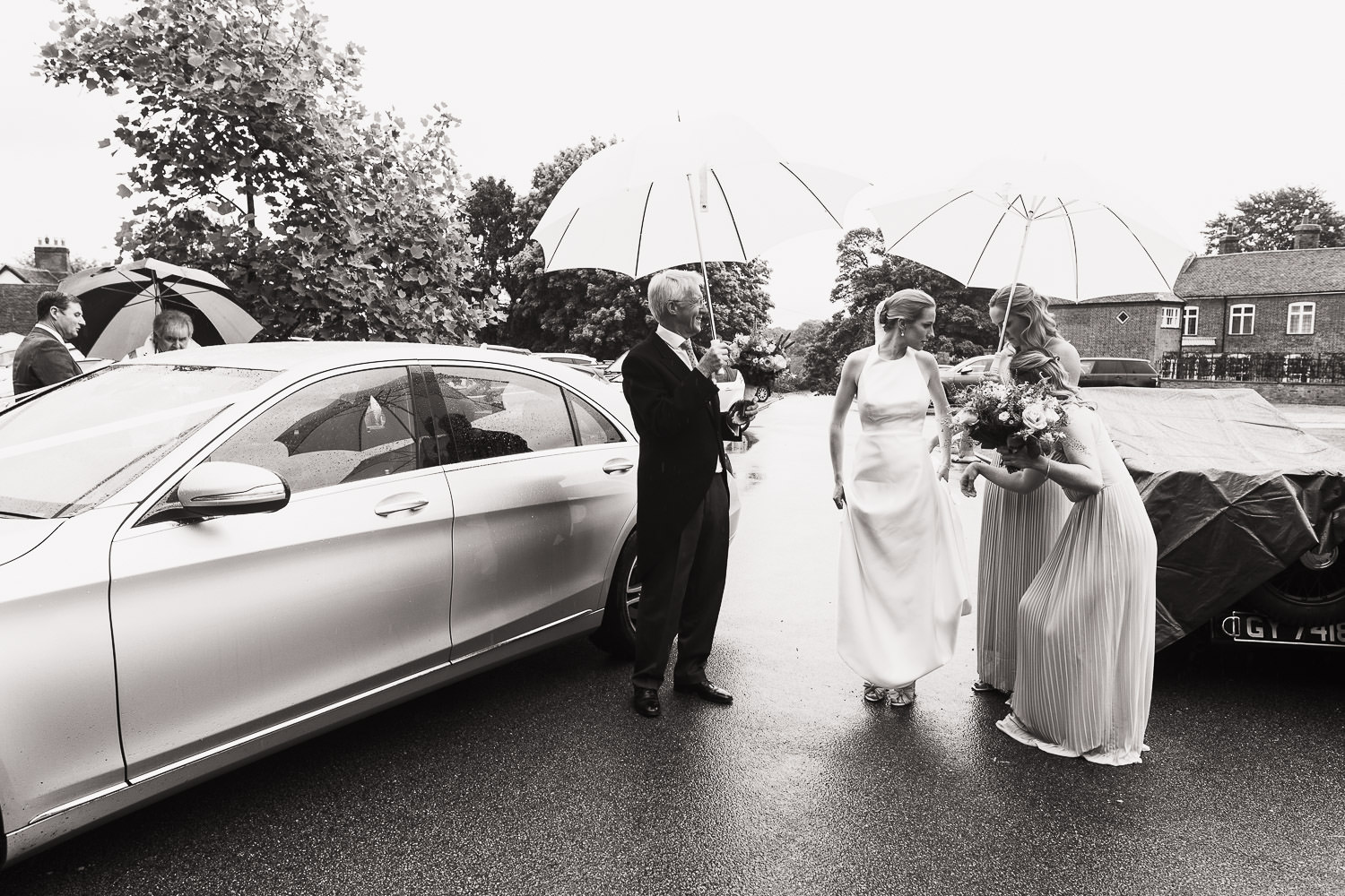 A bride wearing Jesus Peiro 260 dress from Miss Bush is outside the church in Danbury Essex. It is raining and her dad and bridesmaid hold an umbrella. Documentary style Essex wedding photographer.