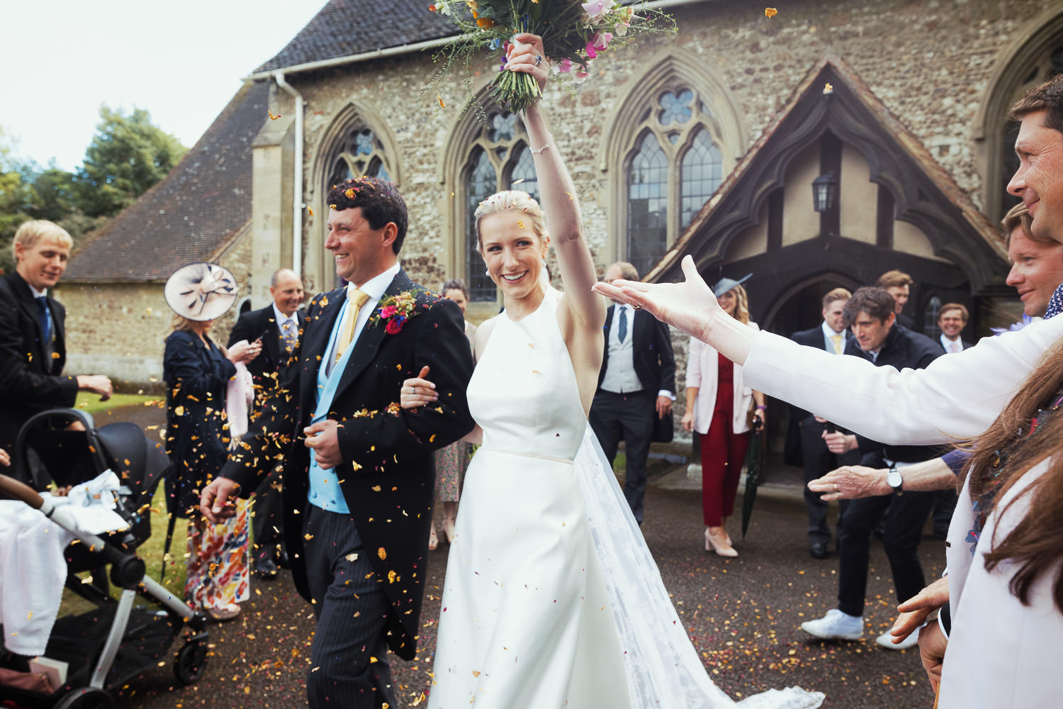 A bride wearing a wedding dress from Miss Bush walks through a shower of confetti with her husband. At Parish Church of St John Baptist in Danbury Essex.