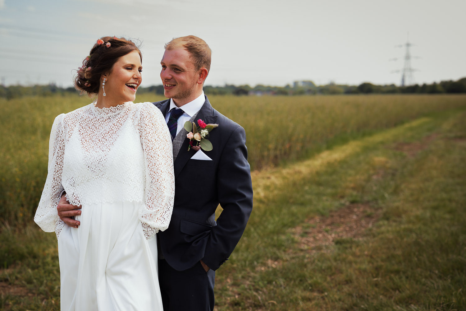 A bride and groom, who are expecting a baby, on their wedding day laughing together in a field in Debden Green.