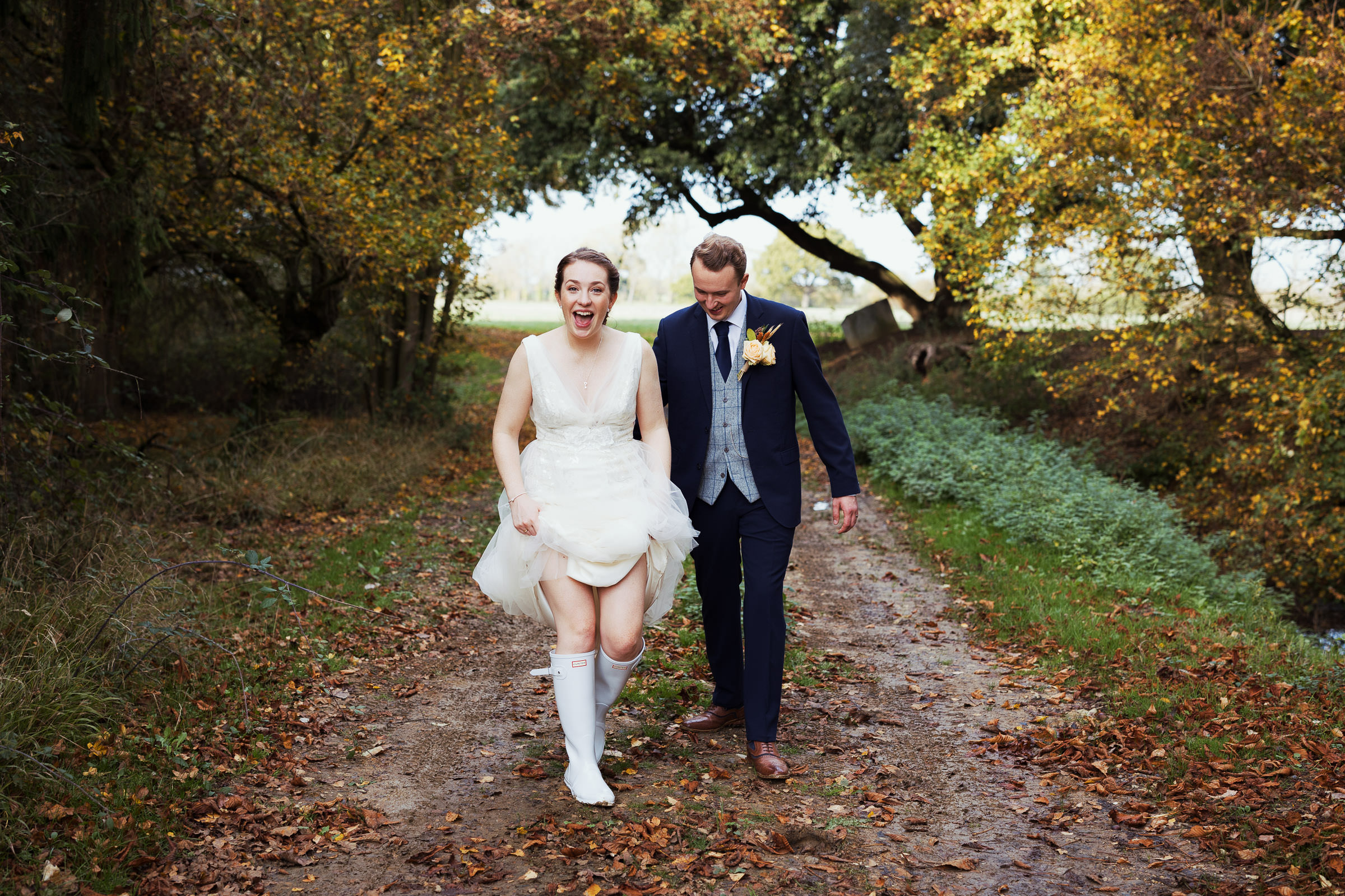 A bride holding her Jenny Packham dress wearing Hunter wellies with her husband. Walking through the leaves at Essex wedding venue Houchins.