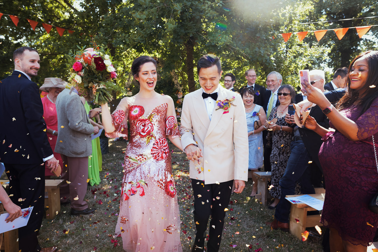 At a garden wedding a bride and groom walk through confetti. The bride is wearing a Temperley carnation sequin gown and the groom is wearing black tie, in a white dinner jacket.