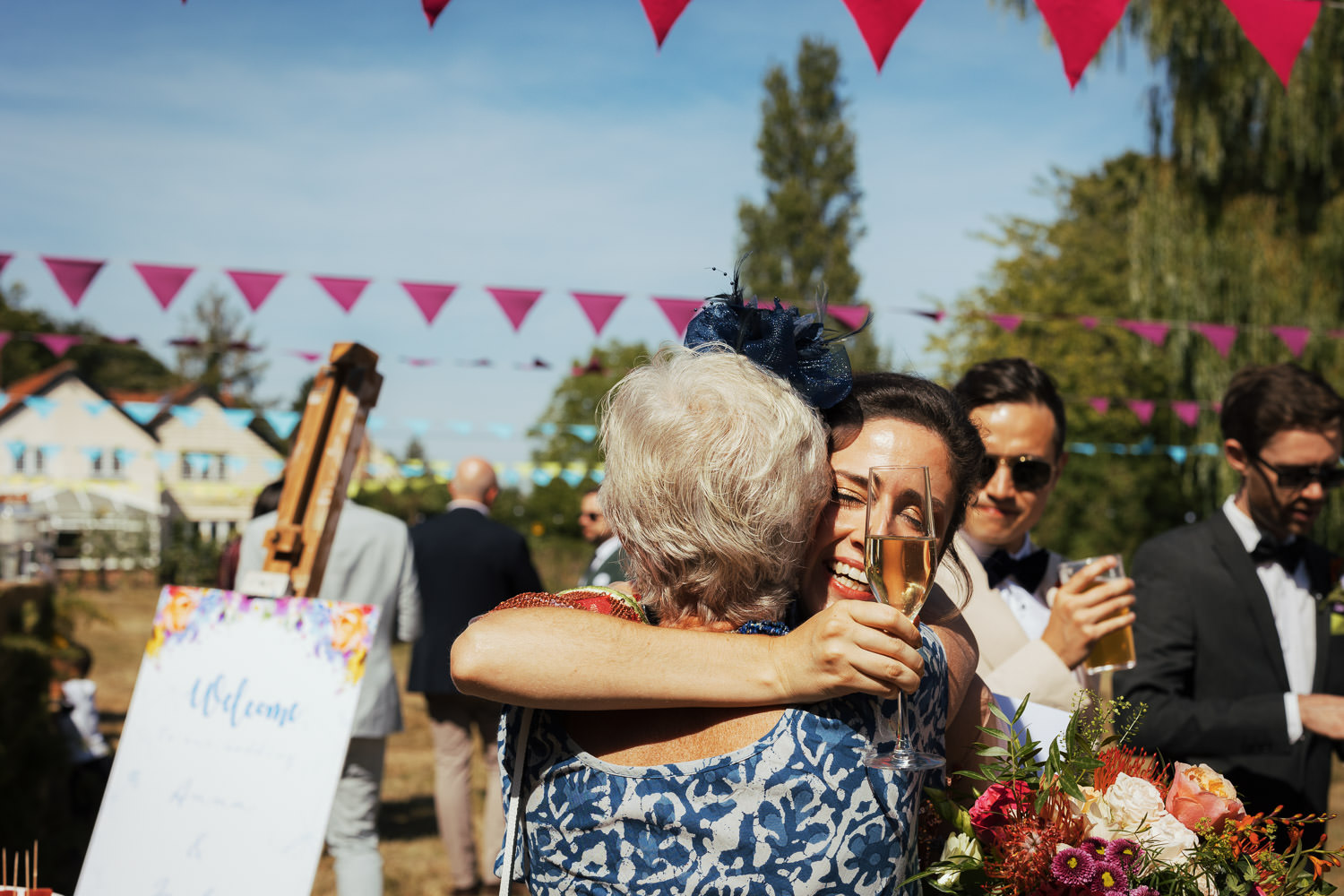 A bride hugs a guest while holding a glass of wine. Under some bunting.