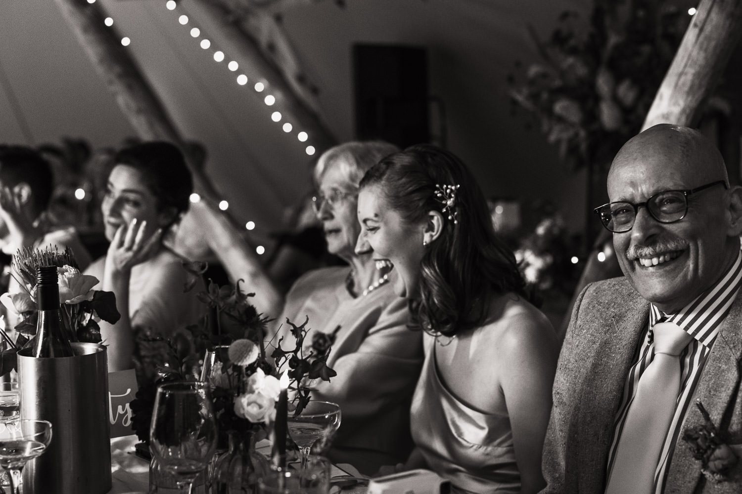 Black and white photo of people sat at a table laughing. At a wedding.
