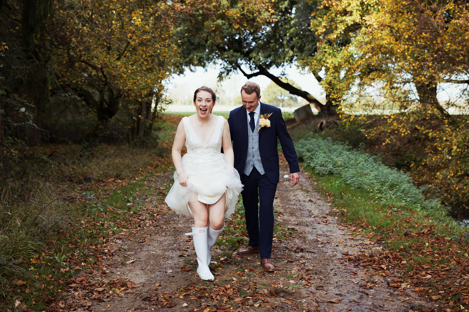 Newly married couple at Houchins in Essex. They are walking along a muddy lane covered with autumnal leaves. The bride is holding up her Jenny Packham dress to reveal her Hunter wellies.