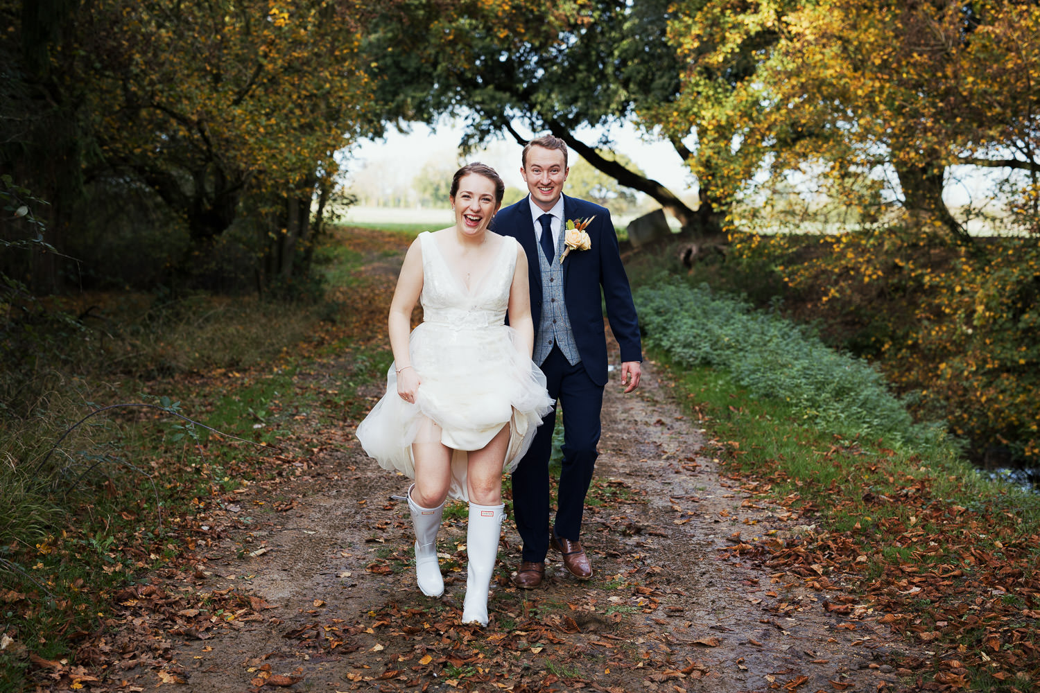 Newly married couple at Houchins in Essex. They are walking along a muddy lane covered with autumnal leaves. The bride is holding up her Jenny Packham dress, May JPB762, to reveal her Hunter wellies.