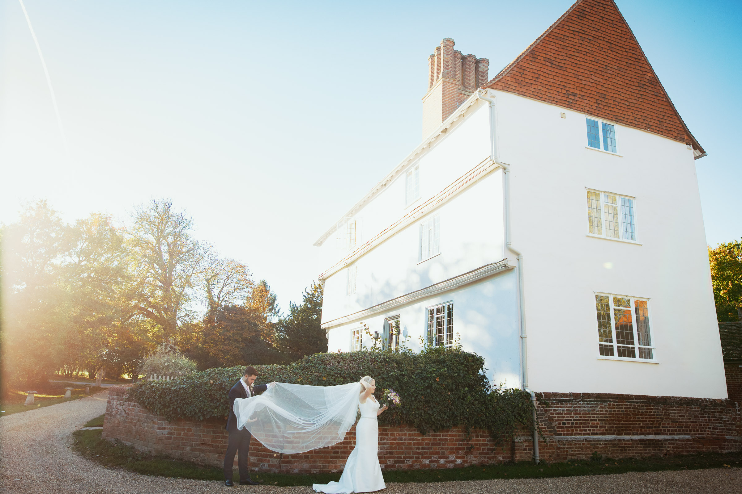 A groom holding his new wife's veil in the wind at a wedding venue in Essex. An autumn wedding photographed by regular Houchins documentary wedding photographer Tracy Morter Photography.