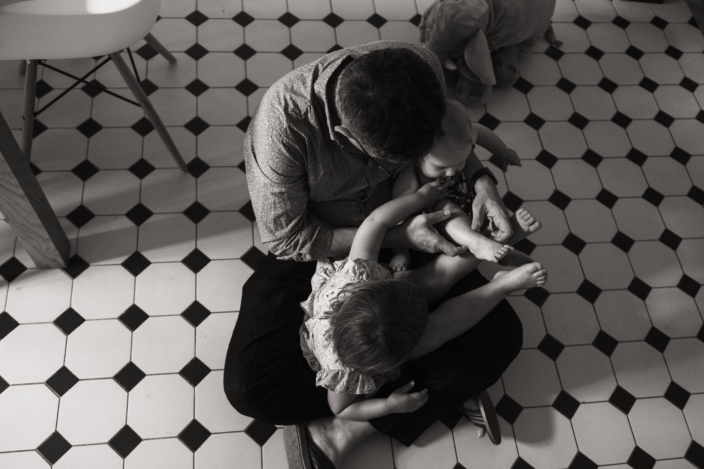 Man sat cross legged on a kitchen floor with his toddler and baby sat on his lap. Documentary family photography in London.