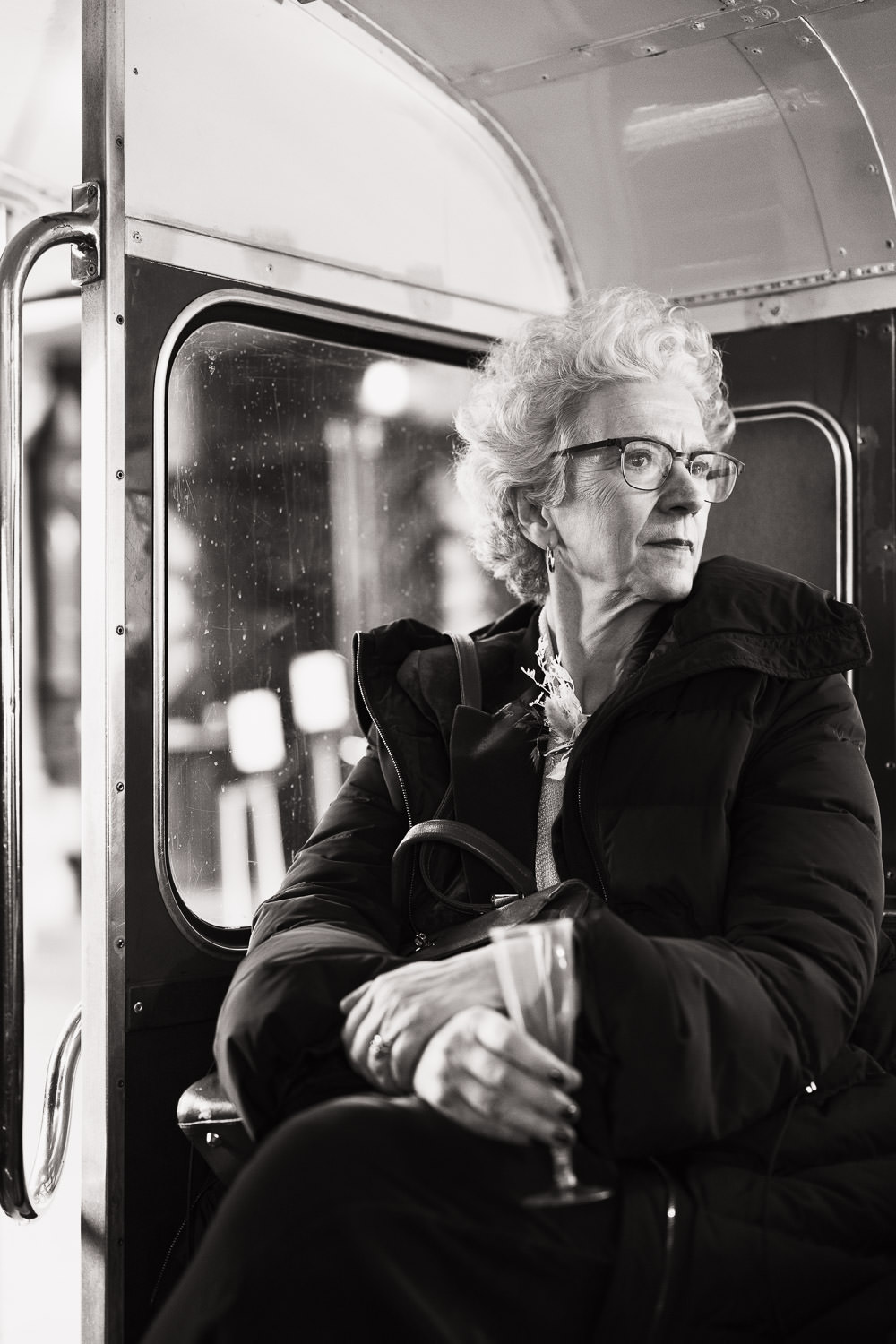 Woman with white hair, in a winter coat, sat on a bus holding a plastic champagne flute on way to wedding reception.