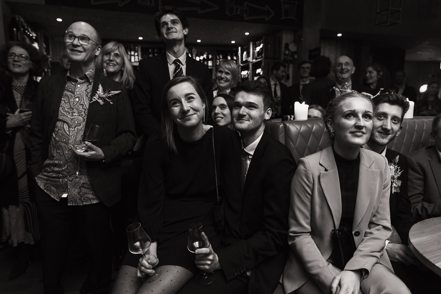 Smiling woman in dotty tights sits on man's lap. Surrounded by guests, they are watching speeches off camera.