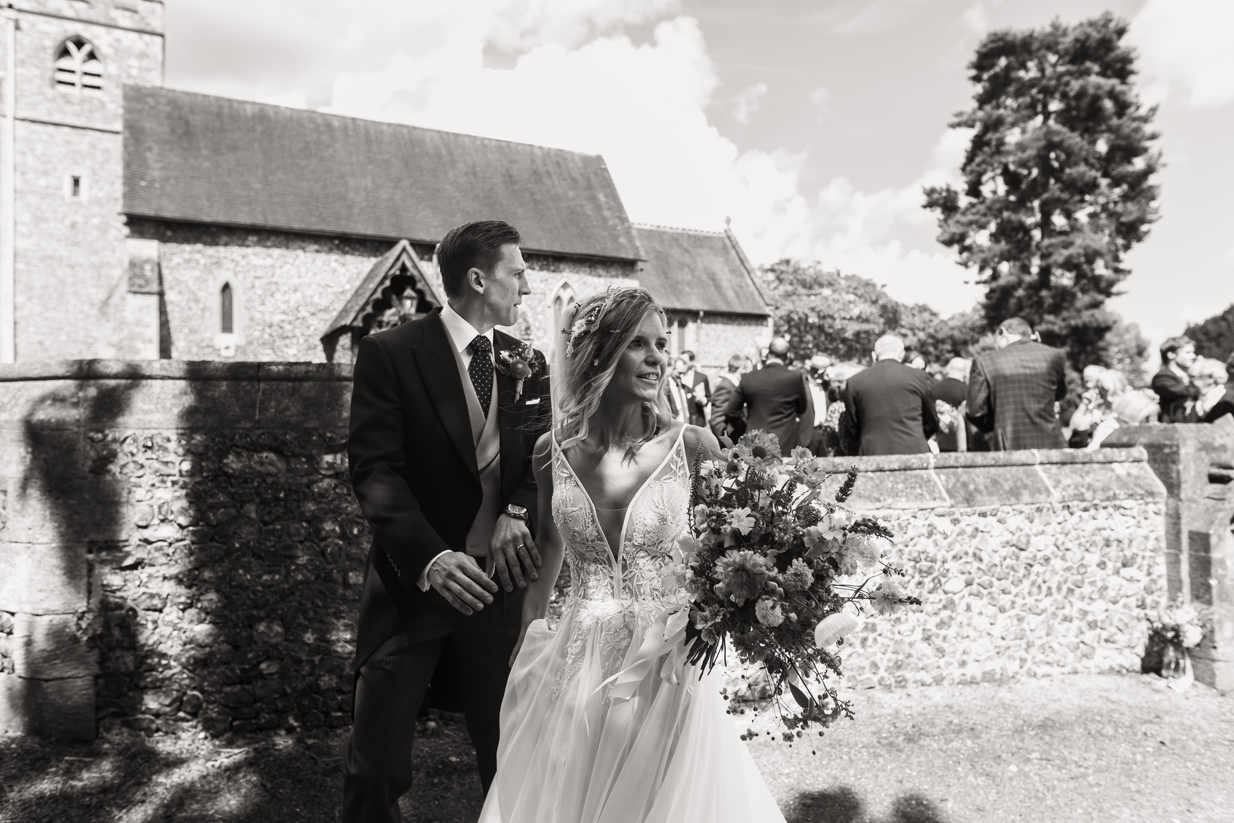 Newly married couple standing outside St Peter and St Paul's church in Shiplake. Wedding guests behind the wall. Documentary style wedding photography.