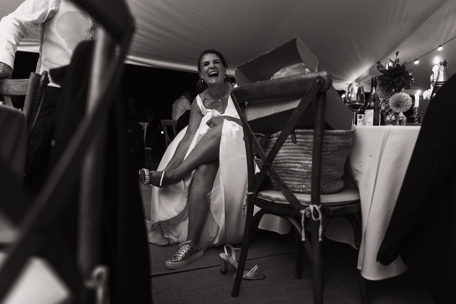 A woman in a wedding dress sits at a table in a marquee from County Marquees Ltd, changing her shoes and laughing. Captured in documentary style wedding photography in Essex at a wedding in Thaxted. Makeup by Sarah Drummond. Featured on a webpage detailing photographer prices and recommending other wedding suppliers near Essex.