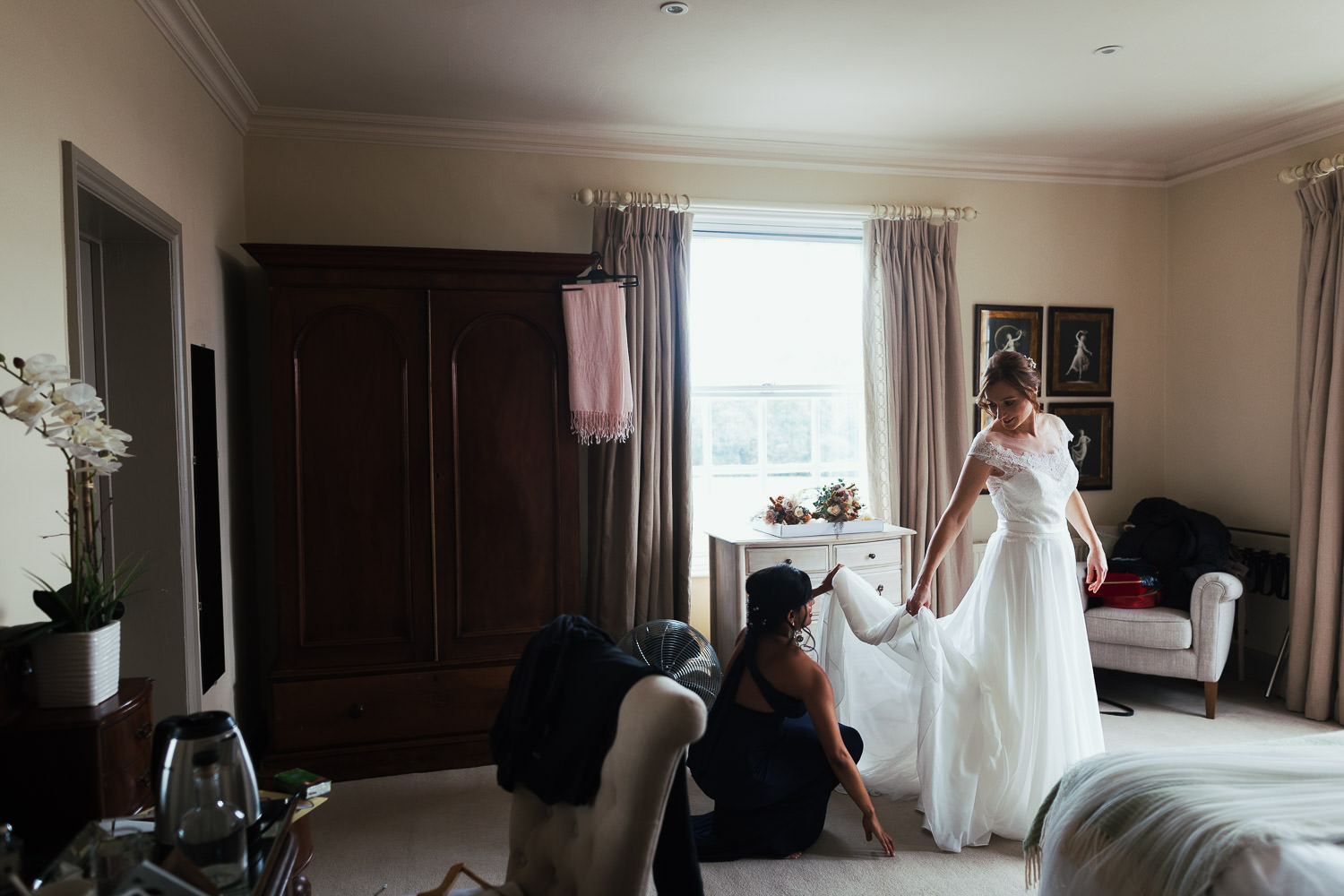 Bridesmaid helping dress bride in hotel in Long Melford, holding the dress up by the window. The Black Lion pub.