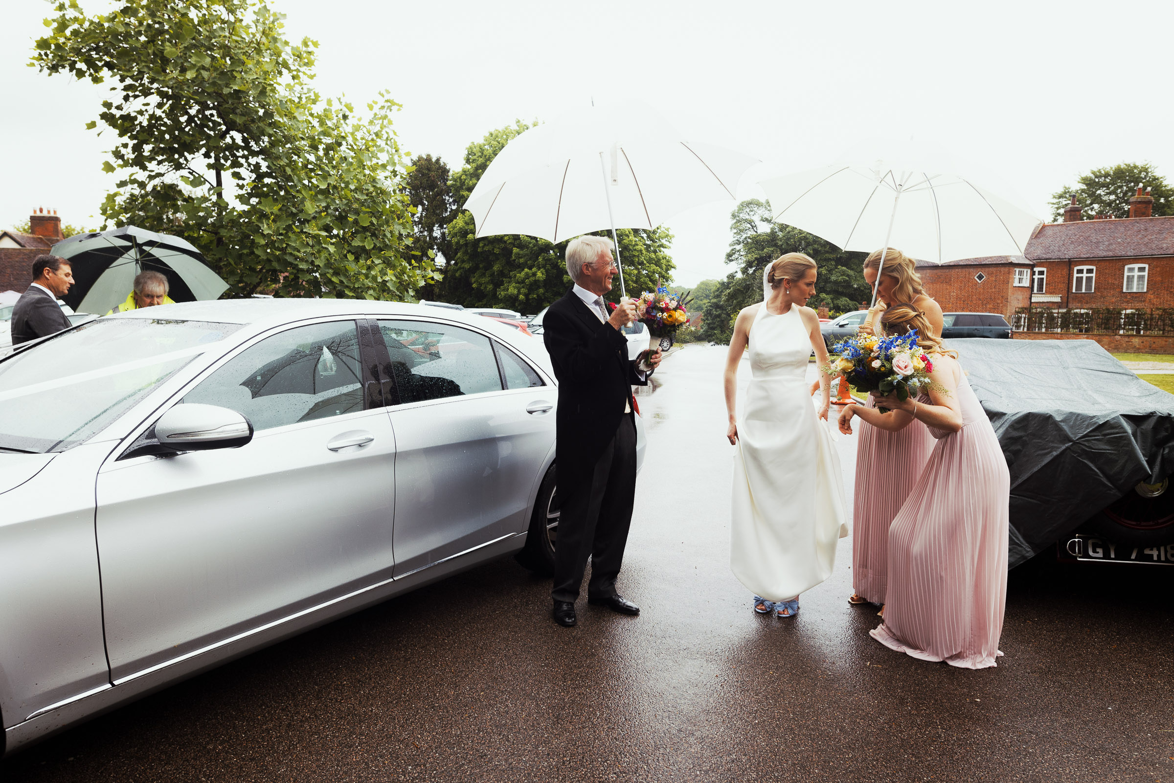 Bride in Jesus Peiro 260 dress from Miss Bush stands outside church in rainy Danbury, Essex. Dad and bridesmaid hold umbrella. Captured by documentary-style Essex wedding photographer from South Woodham Ferrers.