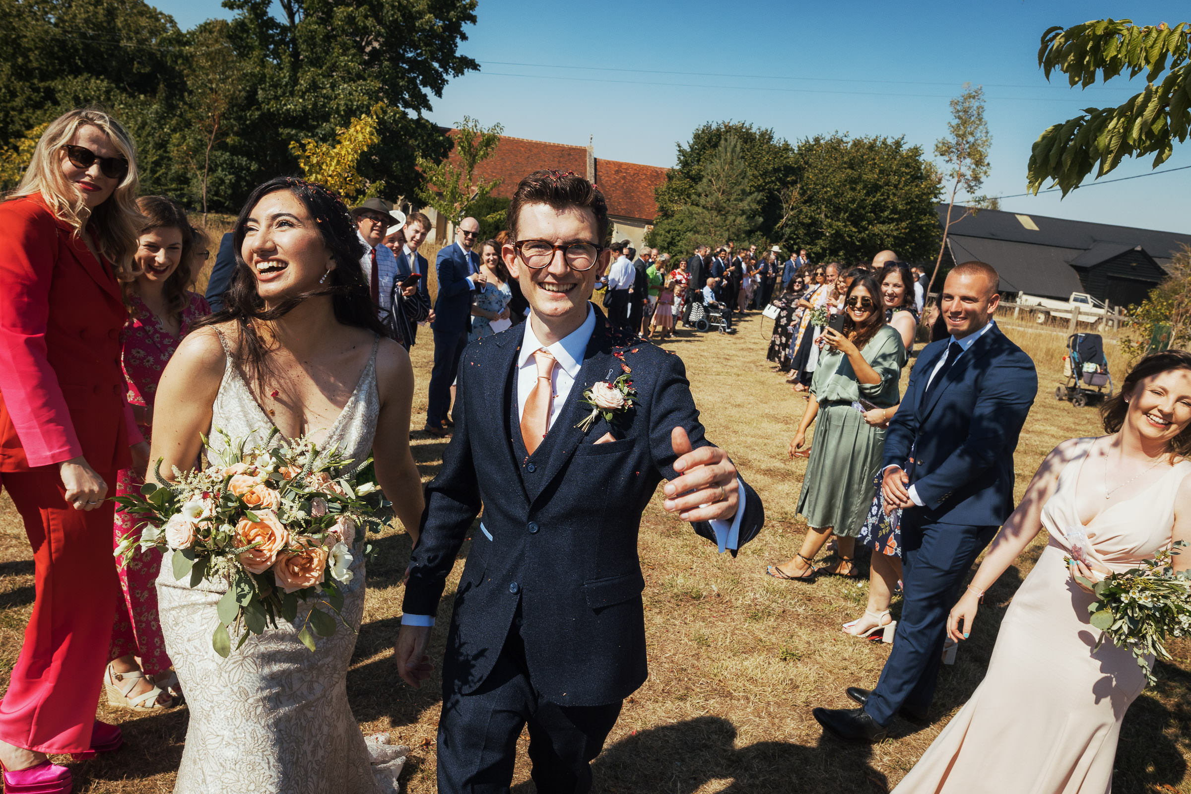 Sunny day and blue skies. At a Suffolk wedding venue called Alpheton Hall Barns a couple walk through confetti thrown by their guests who are in two lines. The bride is laughing.