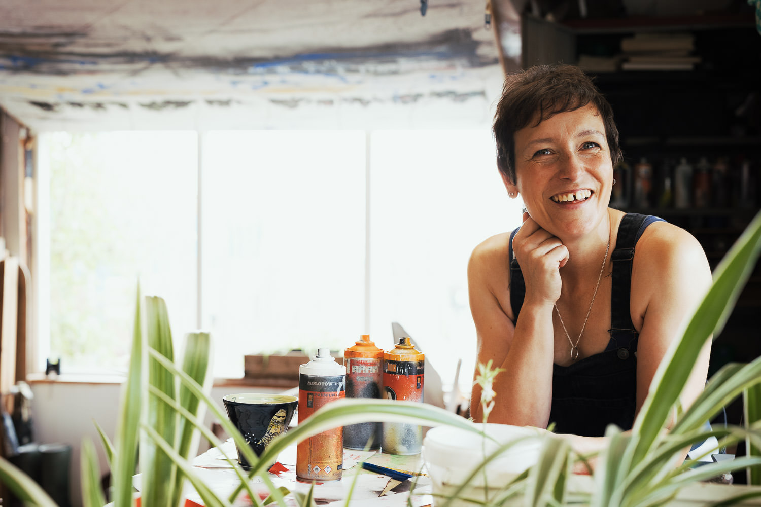 A photograph of Natalie Huntsman aerosol artist in Maldon, Essex. She's leaning on her elbow on a table, in her workshop, spray cans and house plants. Branding photography by Tracy Morter Photography in South Woodham.