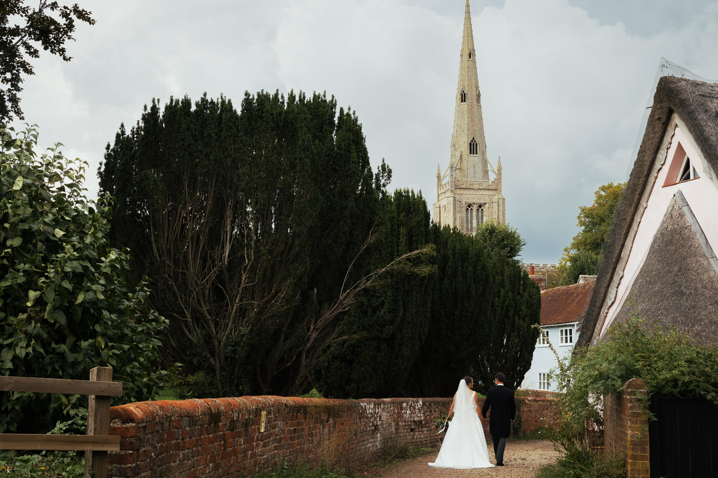 View of newly married couple walking along the lane in Thaxted. The Church of Saint John the Baptist with Our Lady and Saint Laurence in the background, after their wedding.