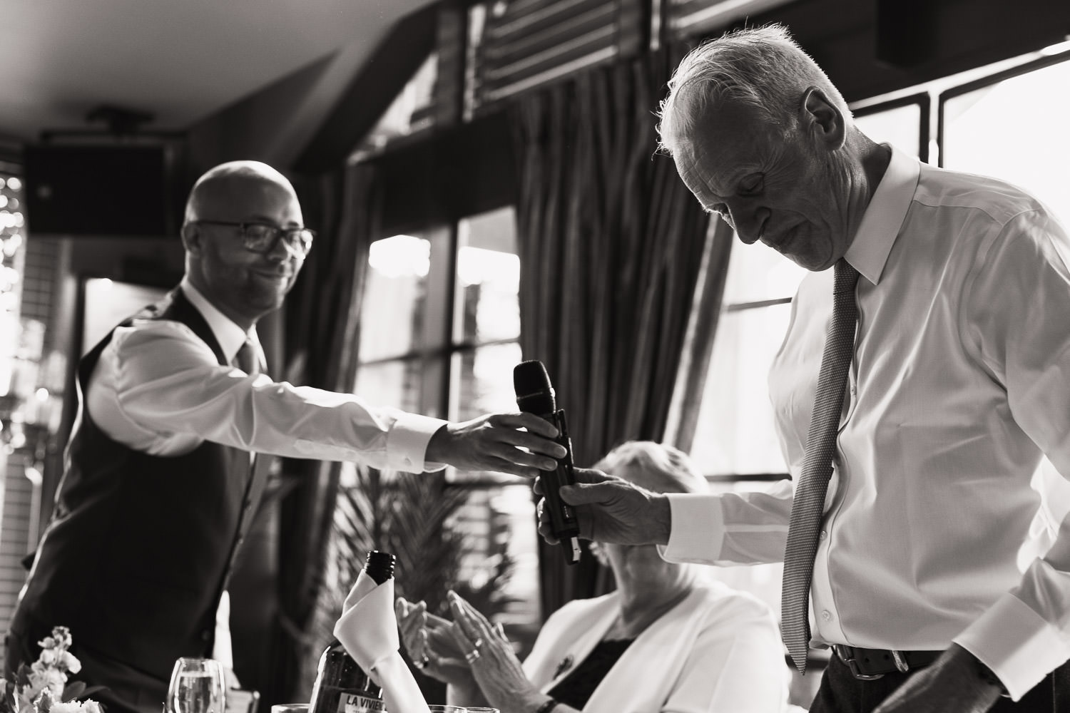 Man in a waistcoat hands microphone to the father of the bride so he can give a speech. Black and white reportage wedding photograph by Chelmsford wedding photographer Tracy Morter.